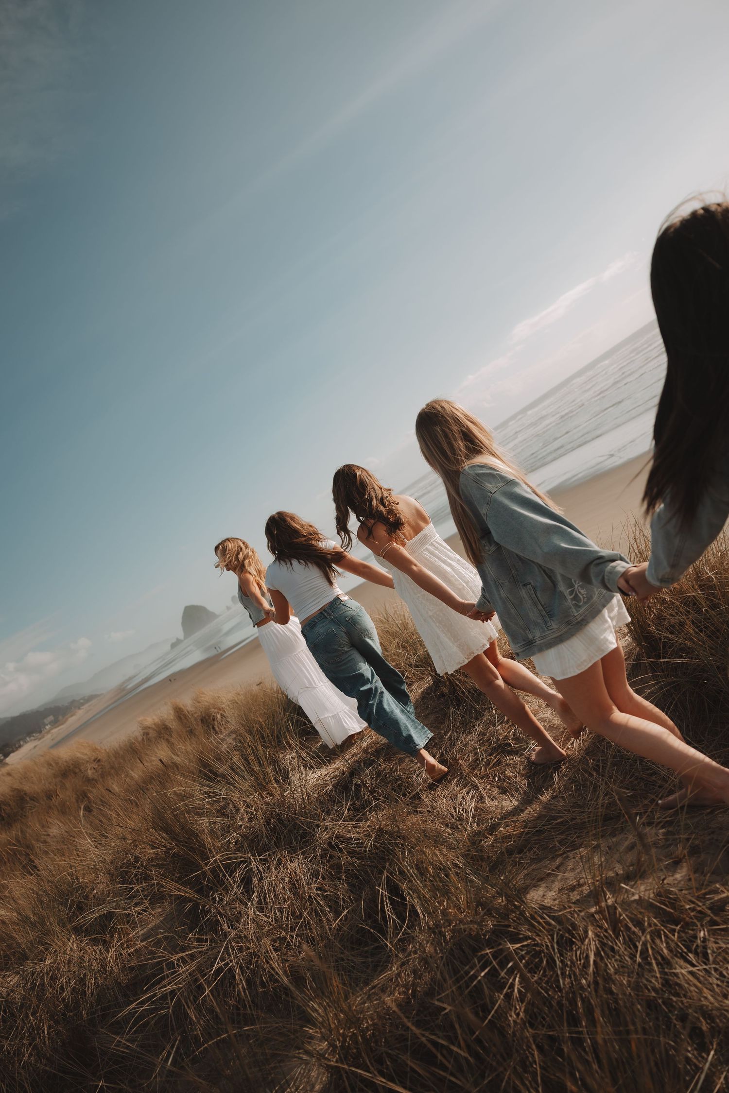 Four young women holding hands walk through golden coastal grass toward the ocean on a sunny day.