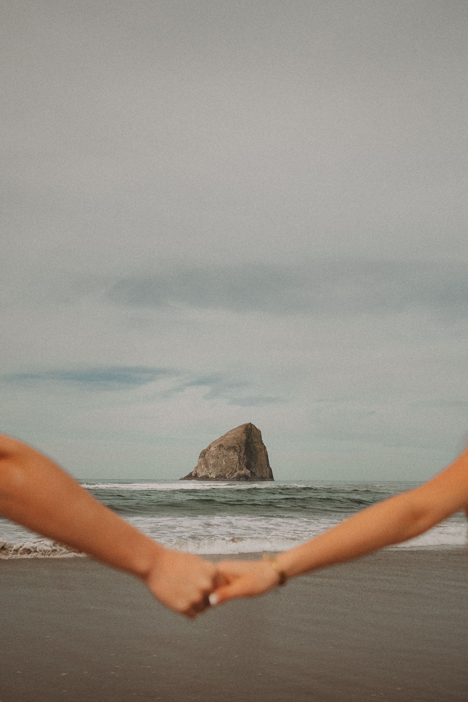Couple holding hands on a beach with a dramatic sea stack rock rising from the ocean in the background.