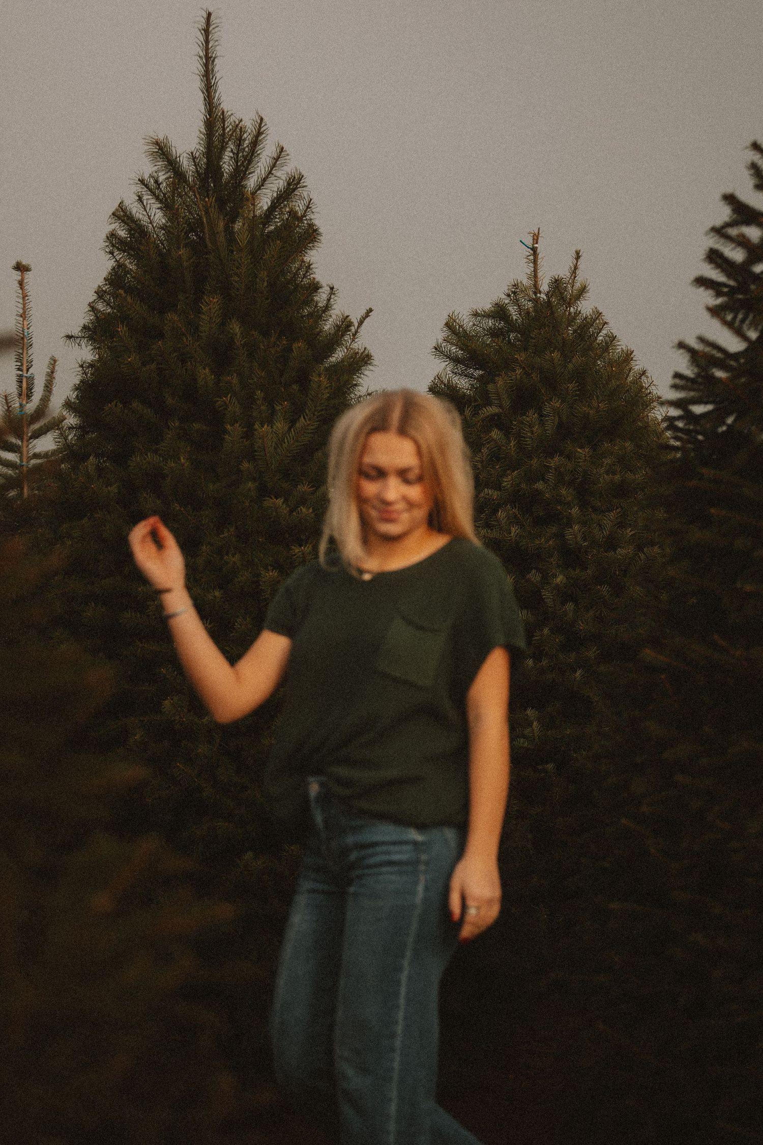 Person in dark green top poses casually against evergreen tree backdrop at dusk.
