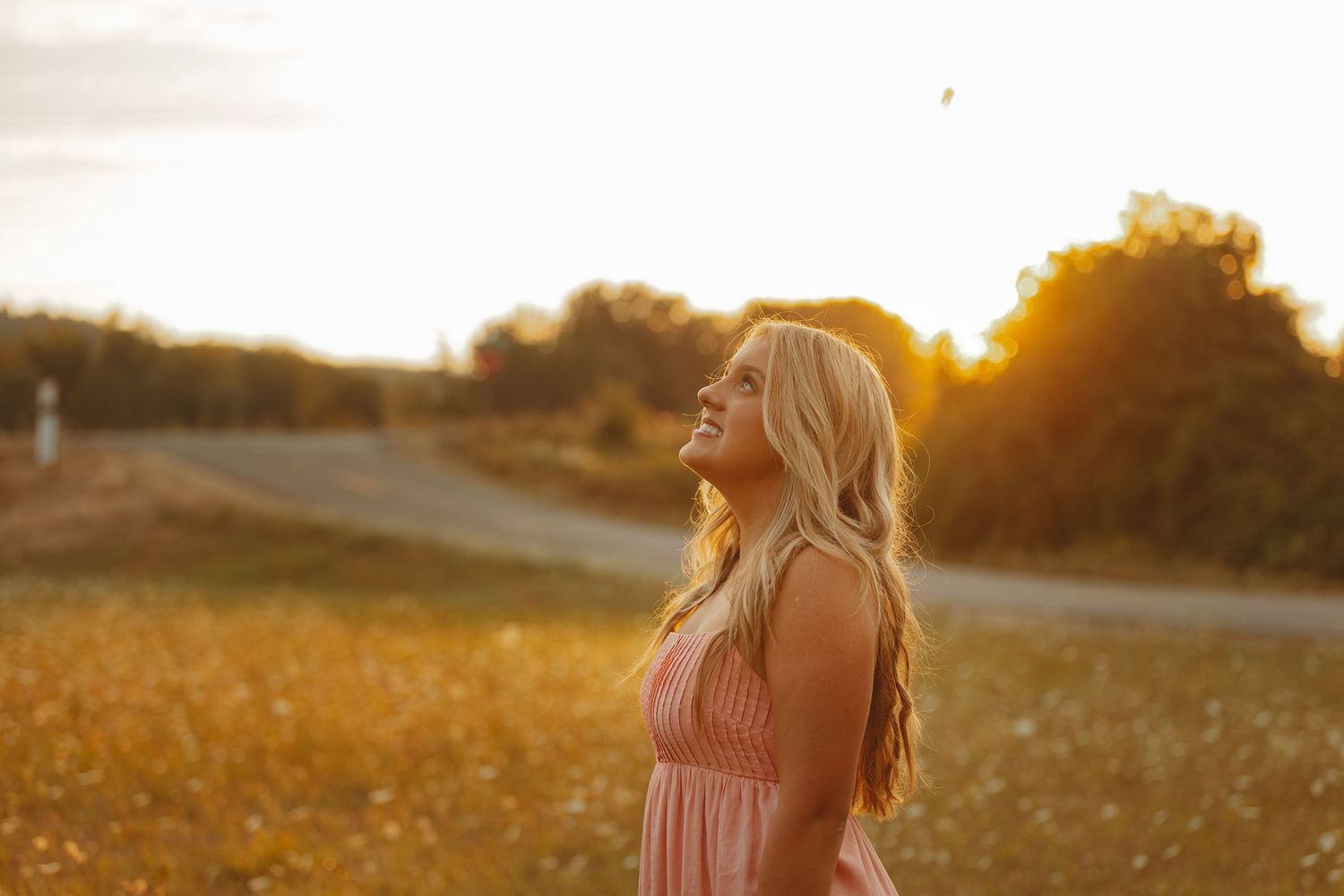Young blonde woman in pink dress standing in golden field at sunset with warm glowing light and trees in background.
