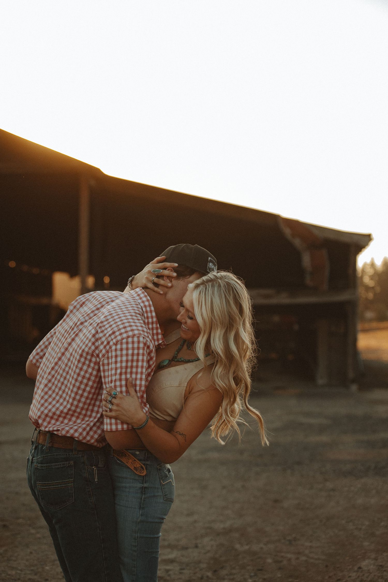 Couple shares tender kiss at golden hour near rustic barn.