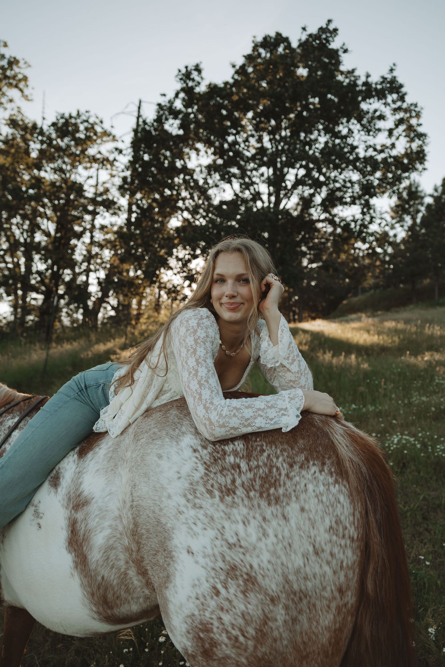 Young blonde woman in white lace top leans on brown and white horse in a sunlit green meadow.