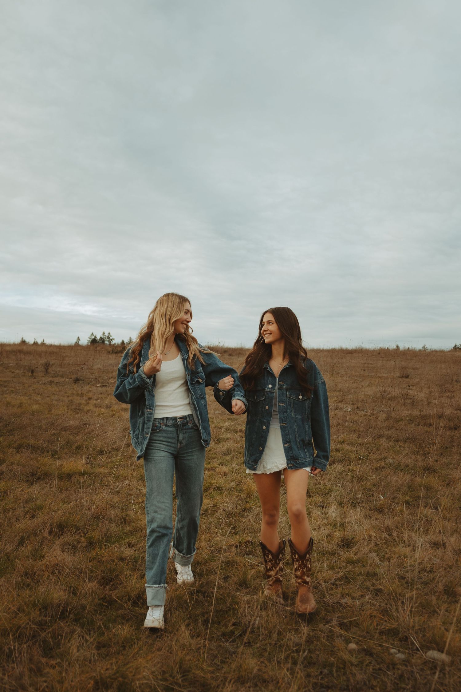 Two friends in denim jackets laugh together while standing in a golden autumn field under cloudy skies.
