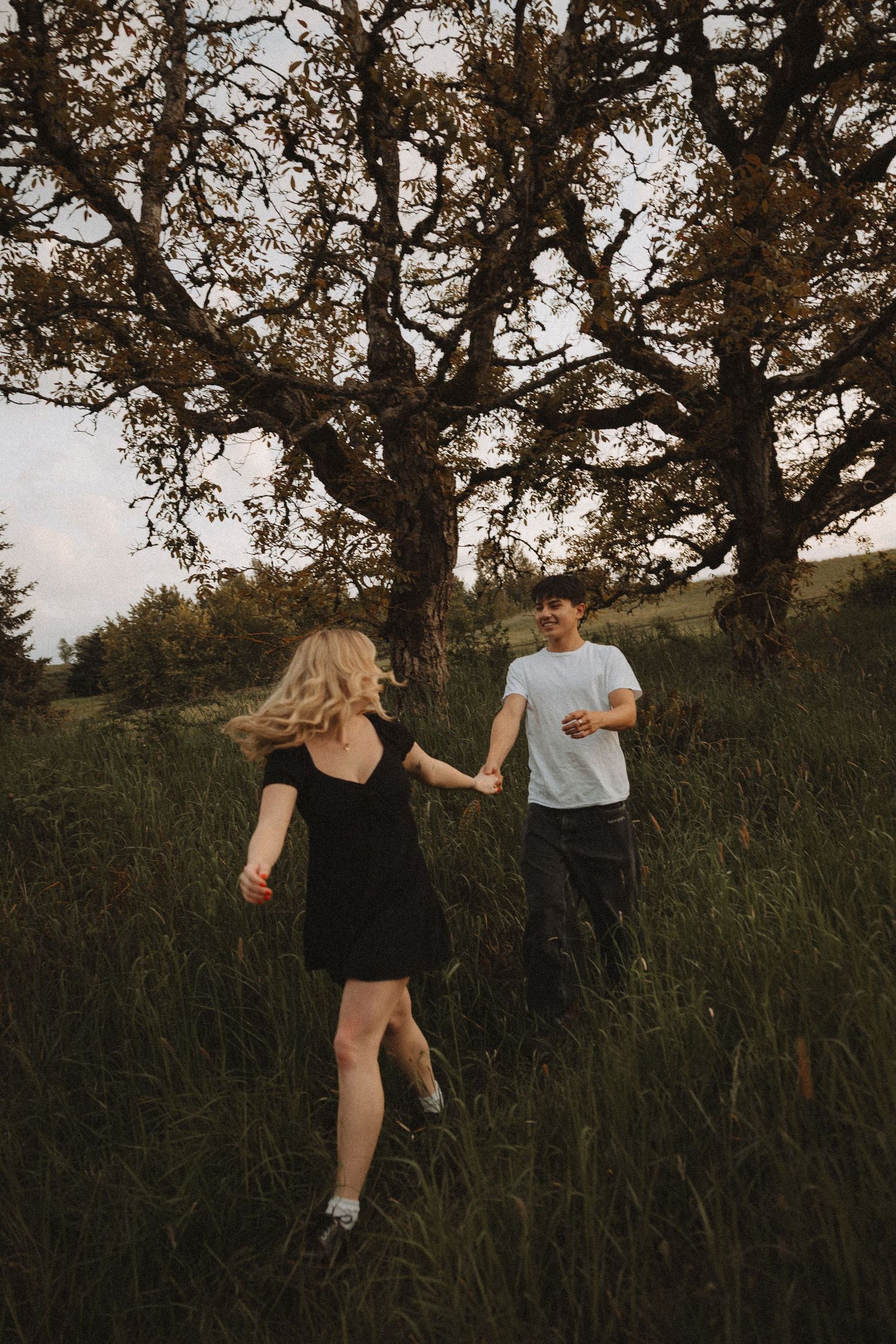Couple holding hands dancing in a lush green meadow beneath a large oak tree during golden hour.