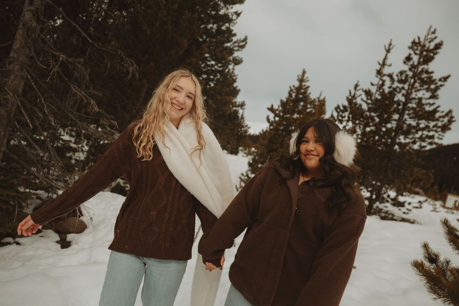 Two women holding hands and smiling in a snowy winter forest wearing brown sweaters and scarves.