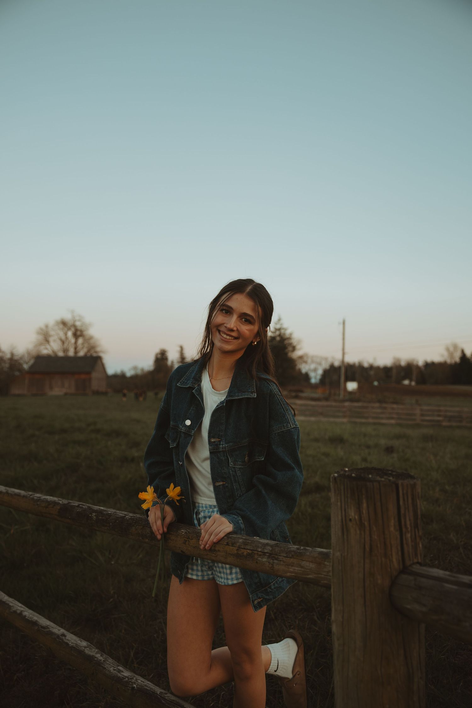 Smiling young woman in denim jacket holds a yellow leaf while leaning on a rustic farm fence.
