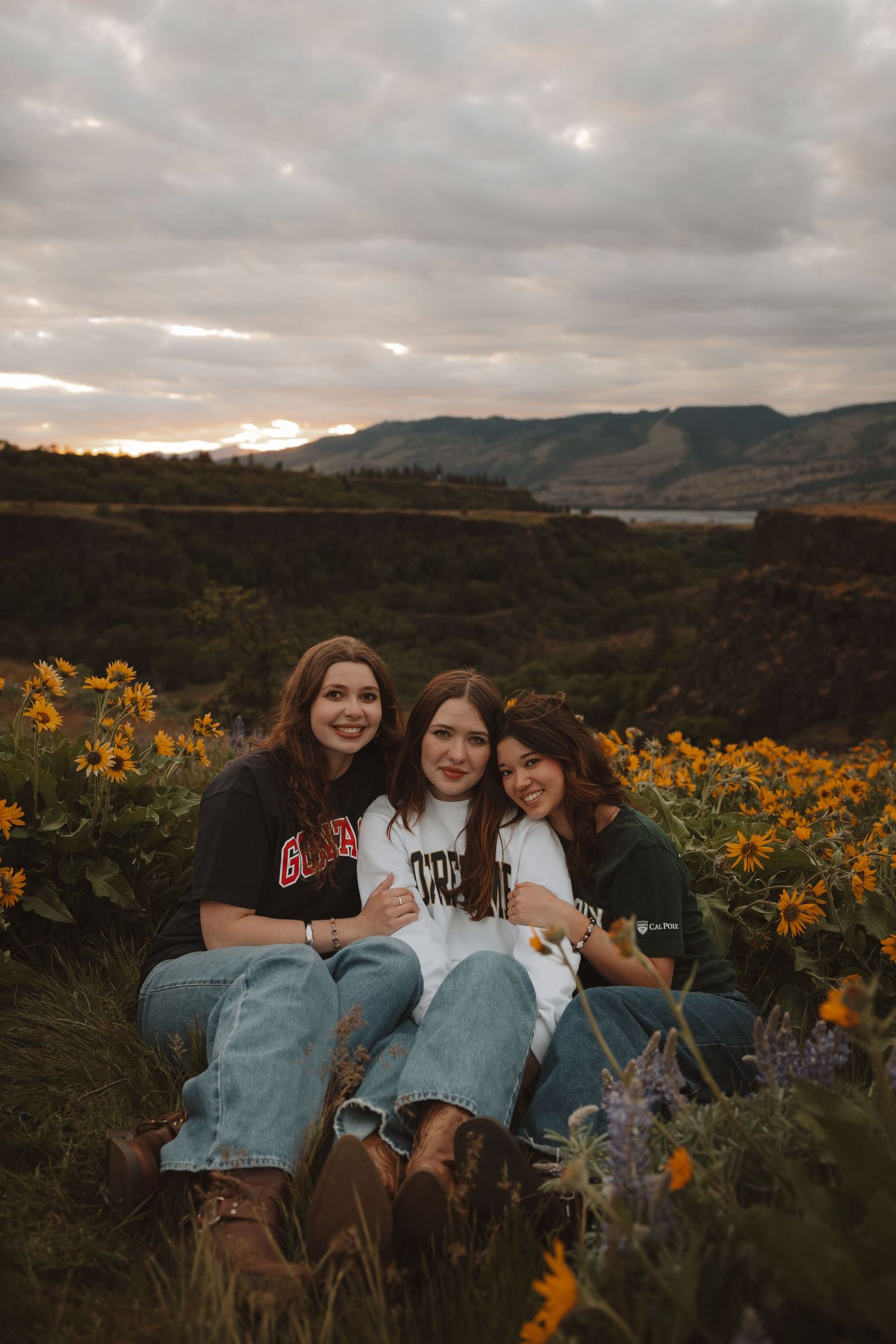 Three young women sitting together in a field of yellow wildflowers at sunset.