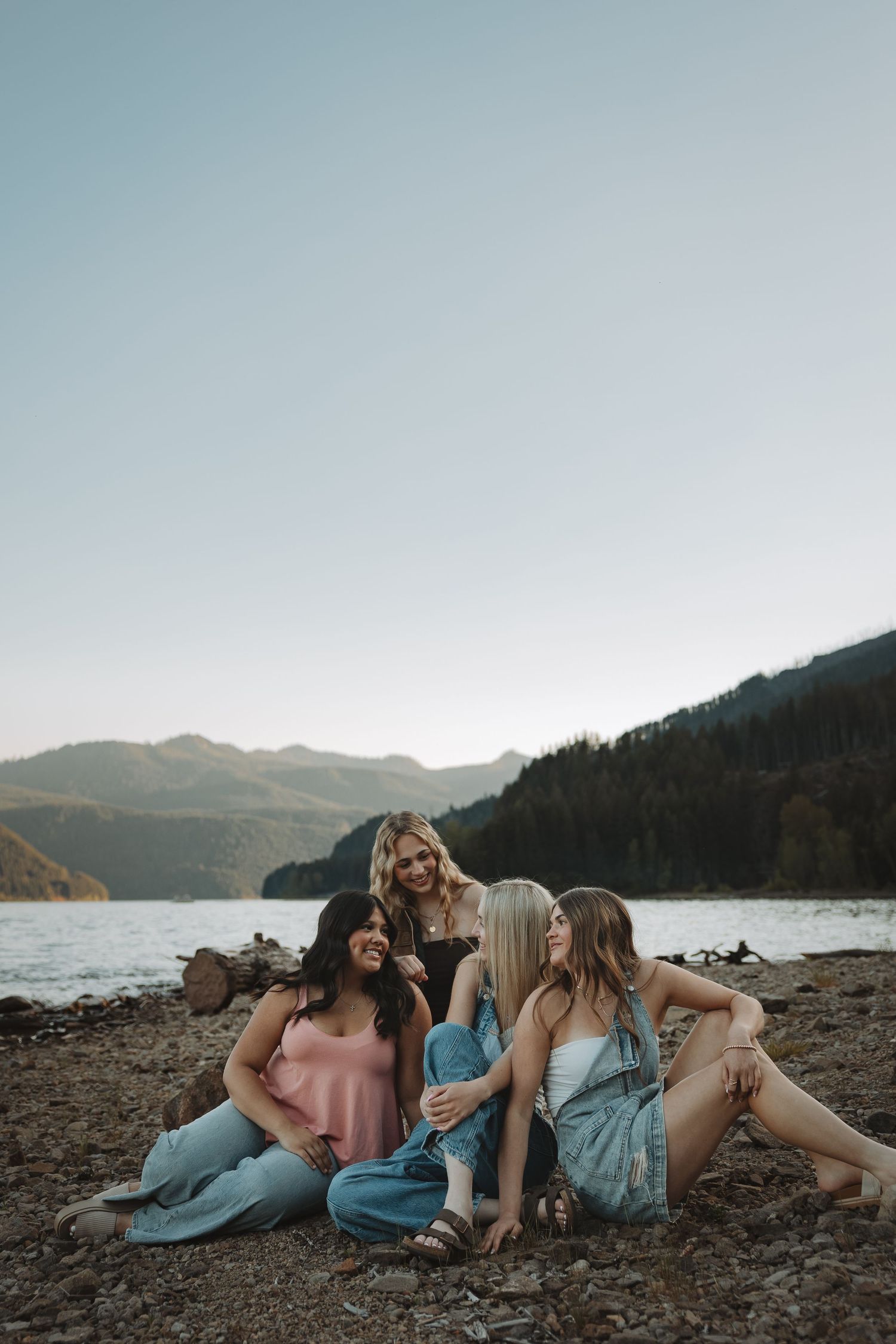 Group of four friends laughing and sitting together on rocky shore near mountain lake at sunset.
