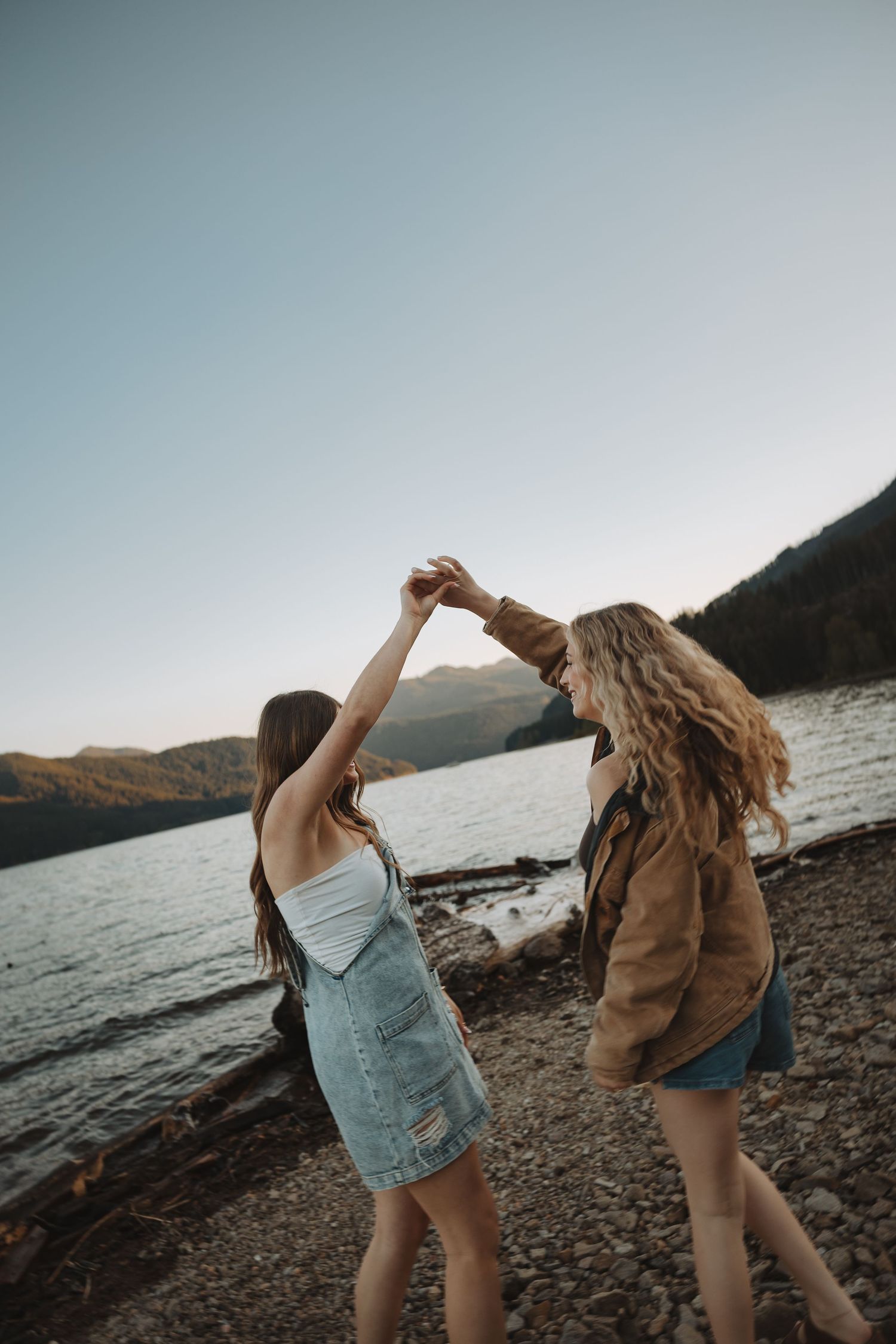 Two young women dancing and holding hands on a rocky shoreline with mountain lake views.