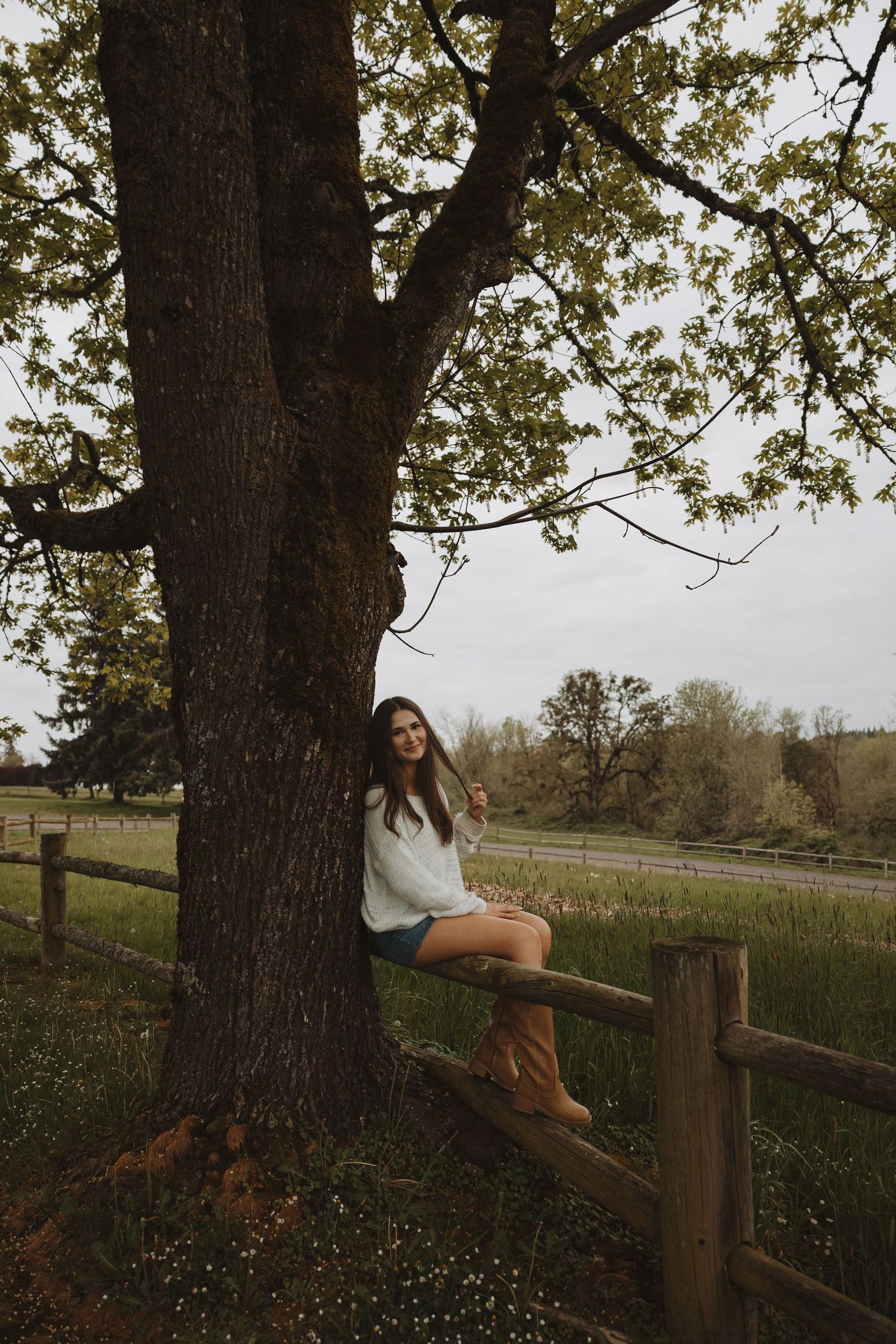 Young woman in white top and boots sitting on wooden fence beside large tree in rural countryside setting.