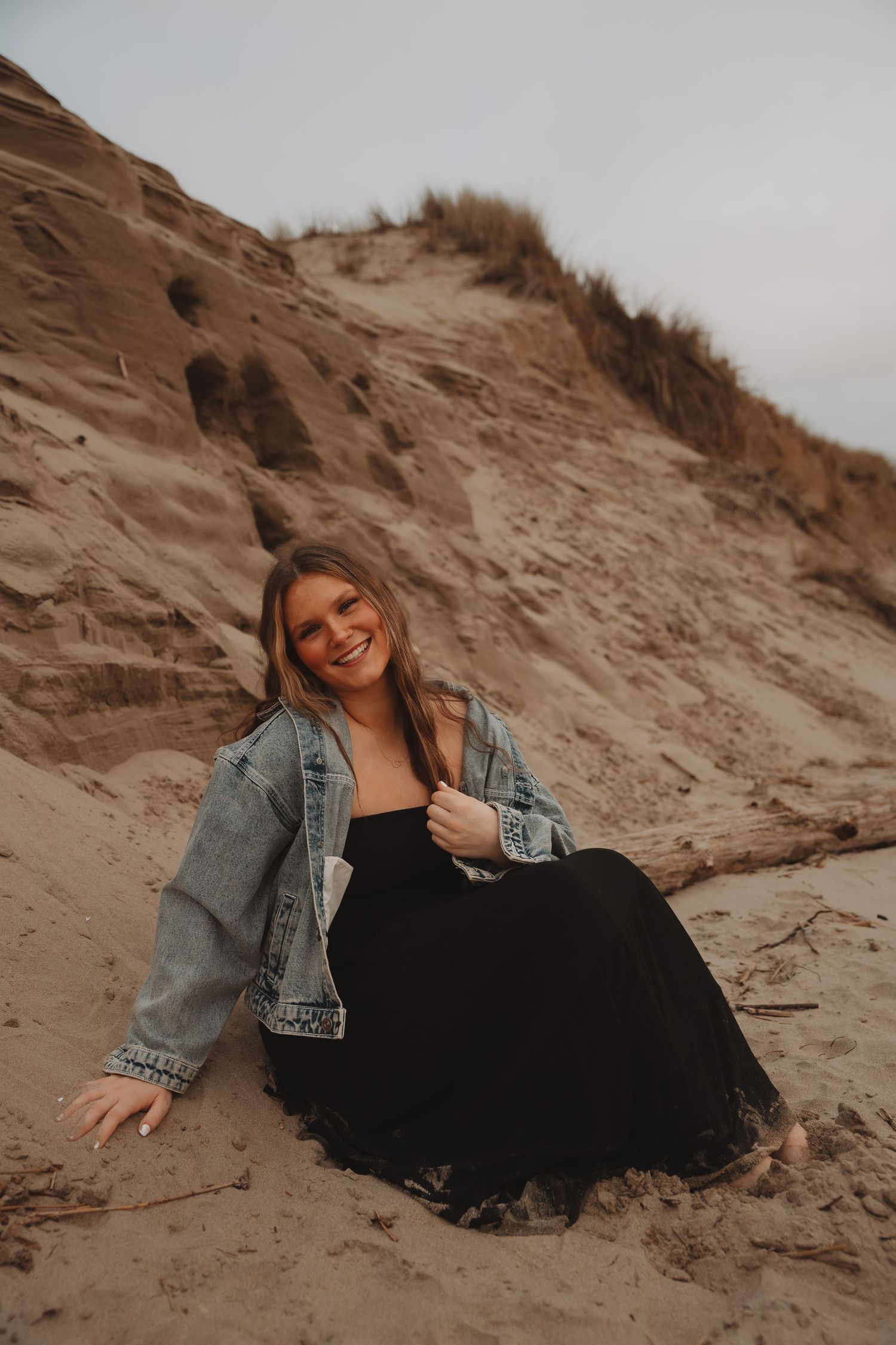 Figure in black dress and denim jacket reclines casually on coastal sand dunes.