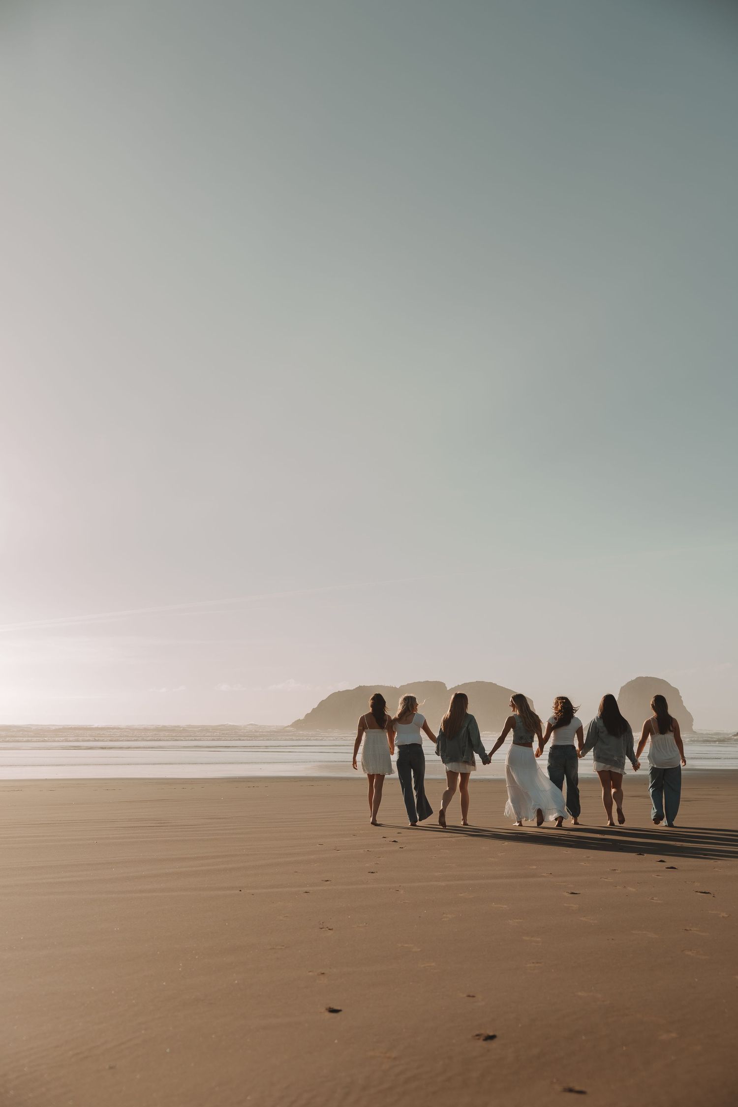 Six women walk hand-in-hand along a sandy beach toward the ocean at golden hour.