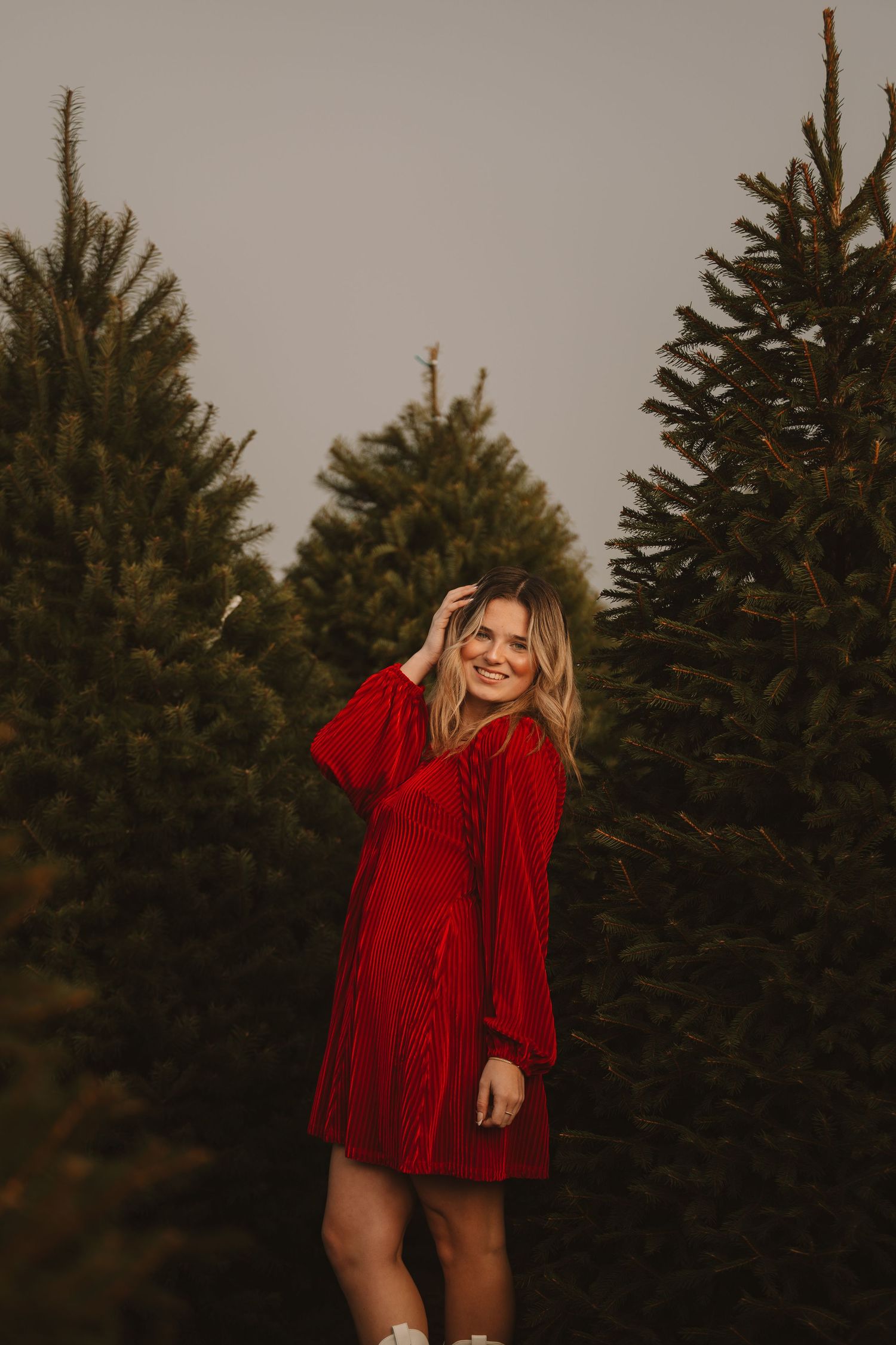 Person in red velvet dress poses expressively against evergreen tree background.