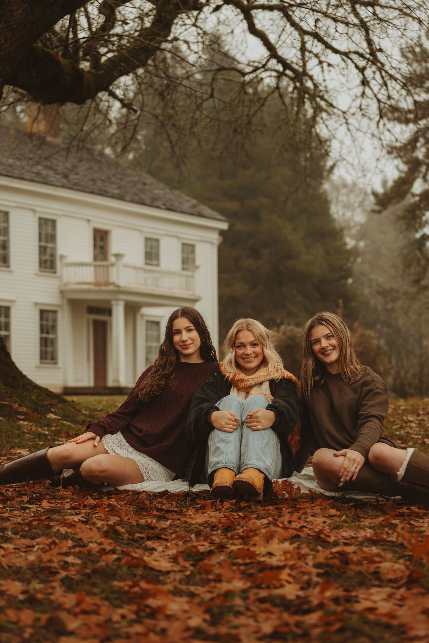 Friends laying in colorful fall leaves wearing cozy sweaters and boots by historic white building.