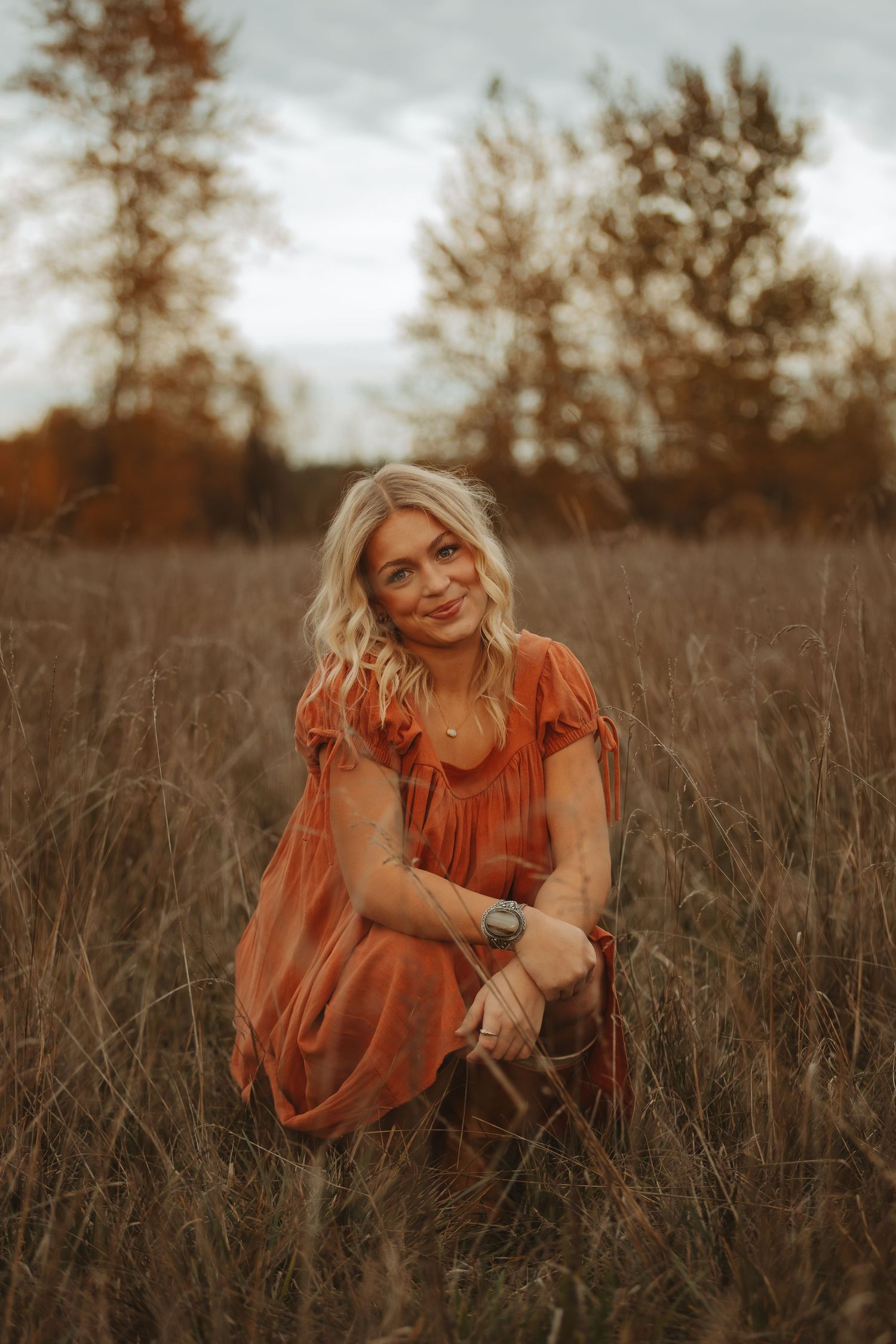 Woman in rust-colored dress poses in tall grass during golden hour autumn photoshoot.
