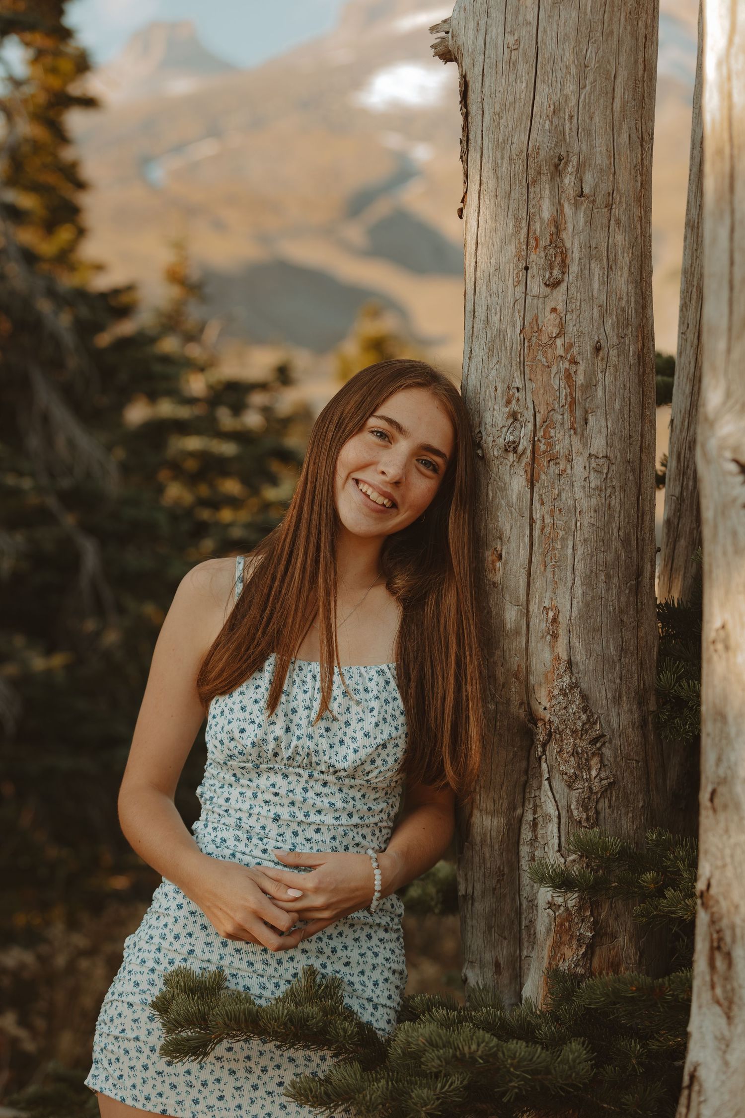 A woman in a floral dress poses against rustic wooden posts during golden hour in a mountain setting.