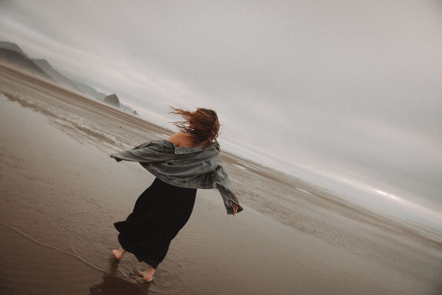 A person in a long dress and sweater twirls joyfully on a misty beach at sunset.