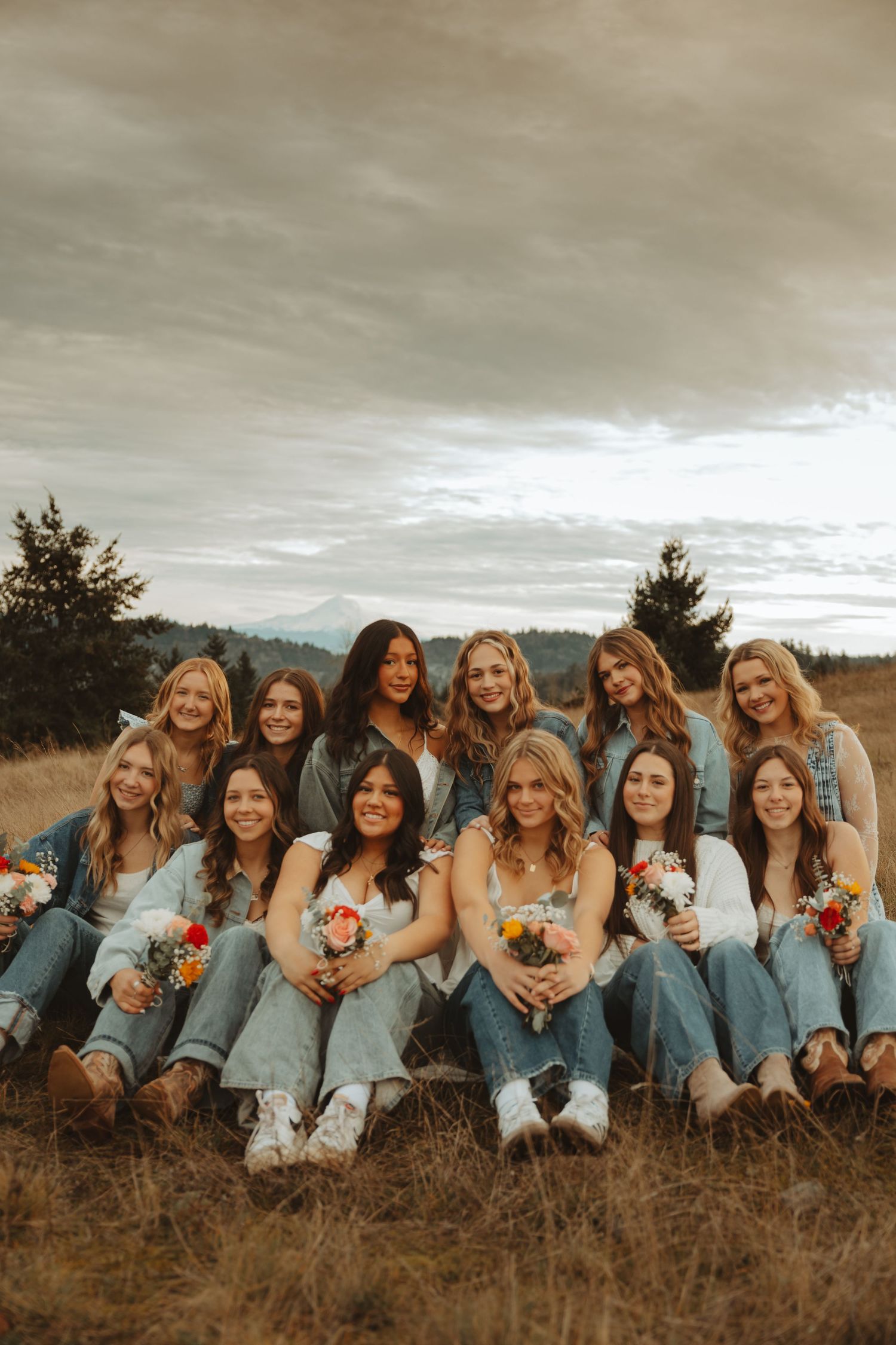A group in matching denim outfits sits together on a hillside holding vibrant floral bouquets.