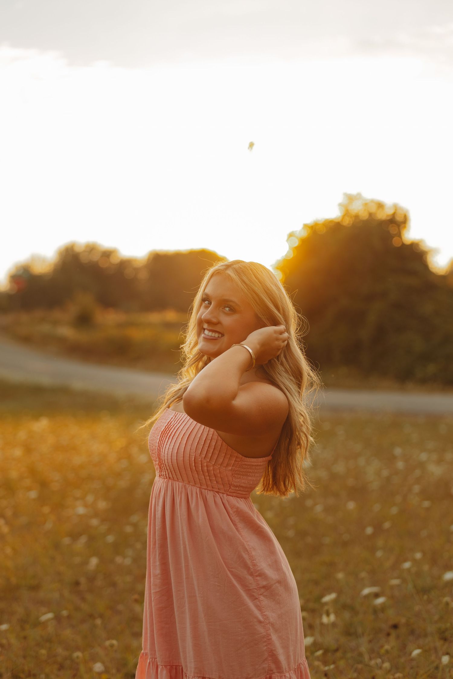 Smiling blonde woman in pink sundress posing in golden meadow during warm sunset light.