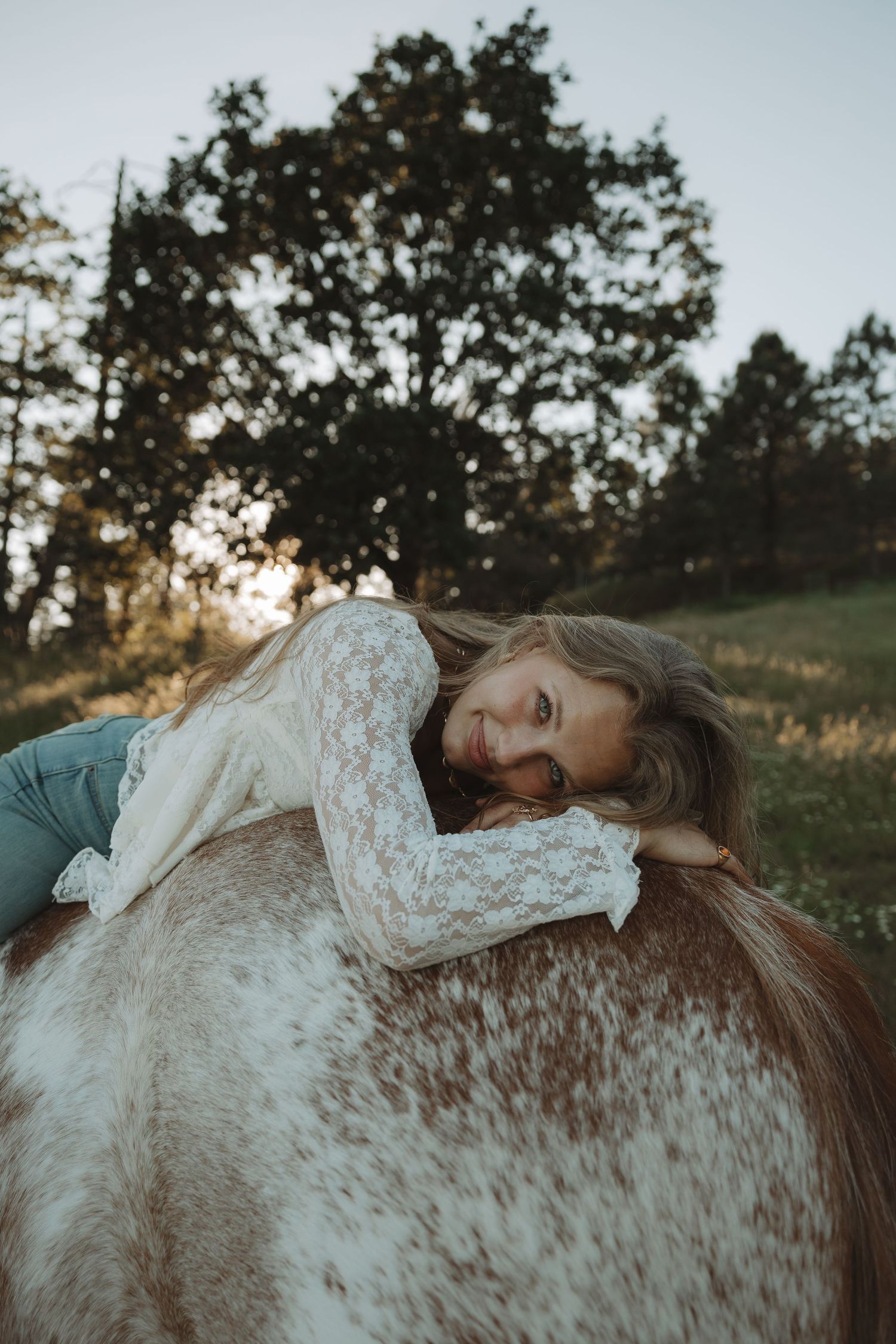 Young woman lying on a horse's back smiling, wearing lace top, golden sunset in background.