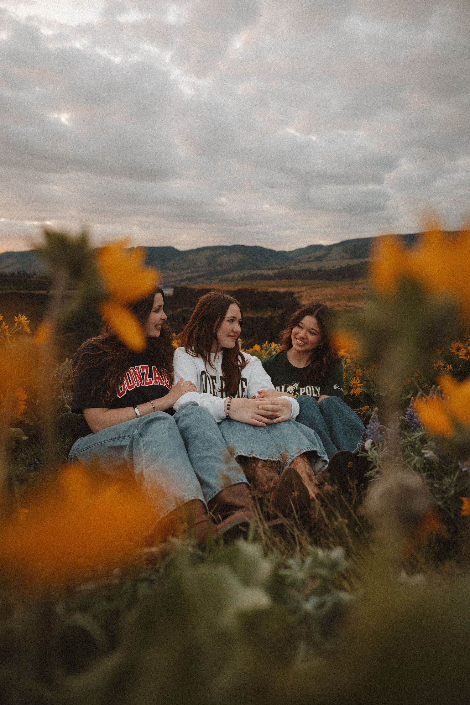 Three young women in college sweatshirts laugh and chat while sitting in a yellow wildflower field at sunset.