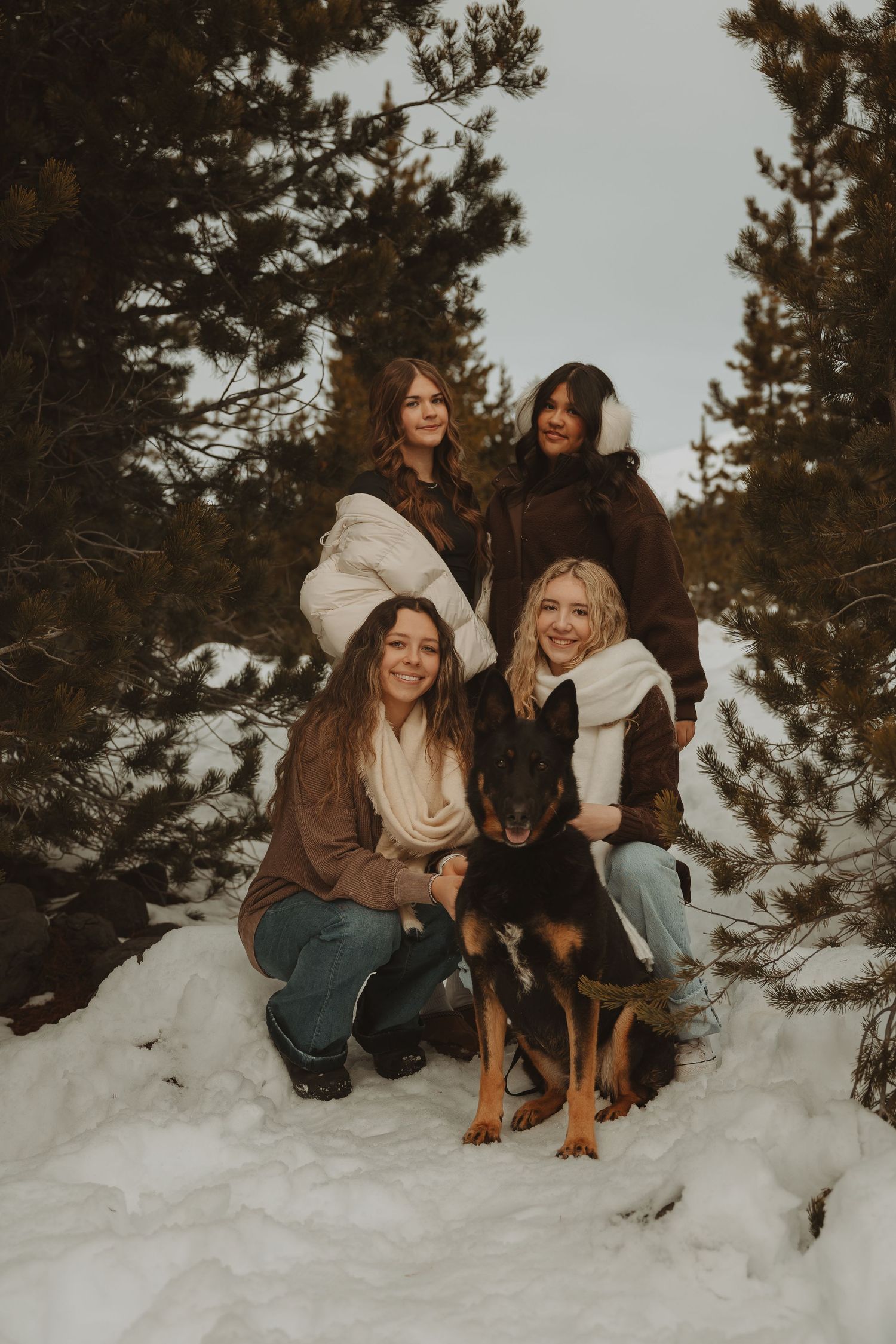 Four women posing with a German Shepherd dog in a snowy pine forest during winter.