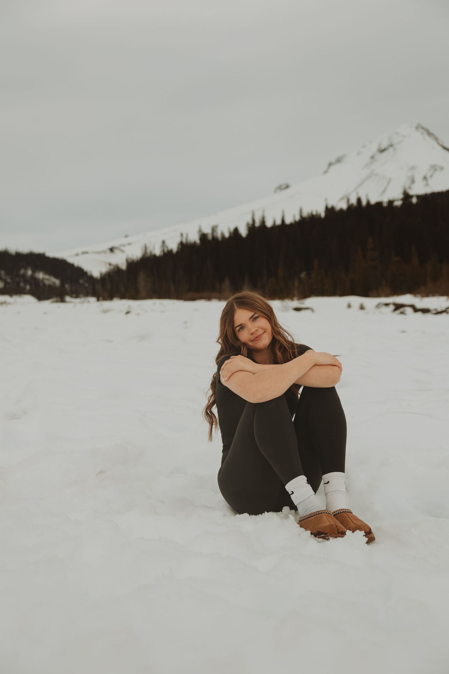 Young woman in black outfit sits cross-legged on snow with scenic mountain and pine forest backdrop.