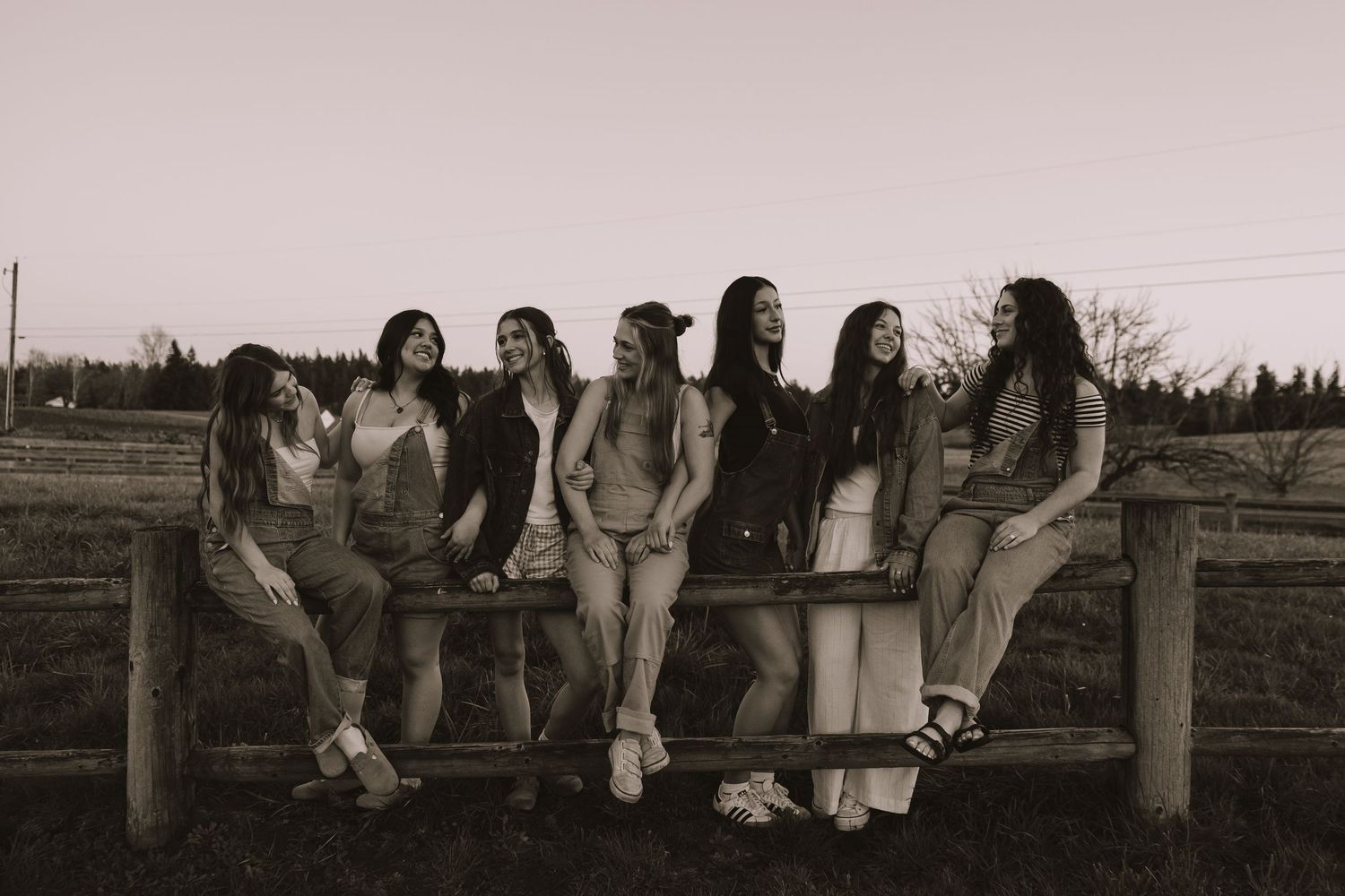 Group of seven girls laughing together leaning on a fence at dusk in sepia tone.