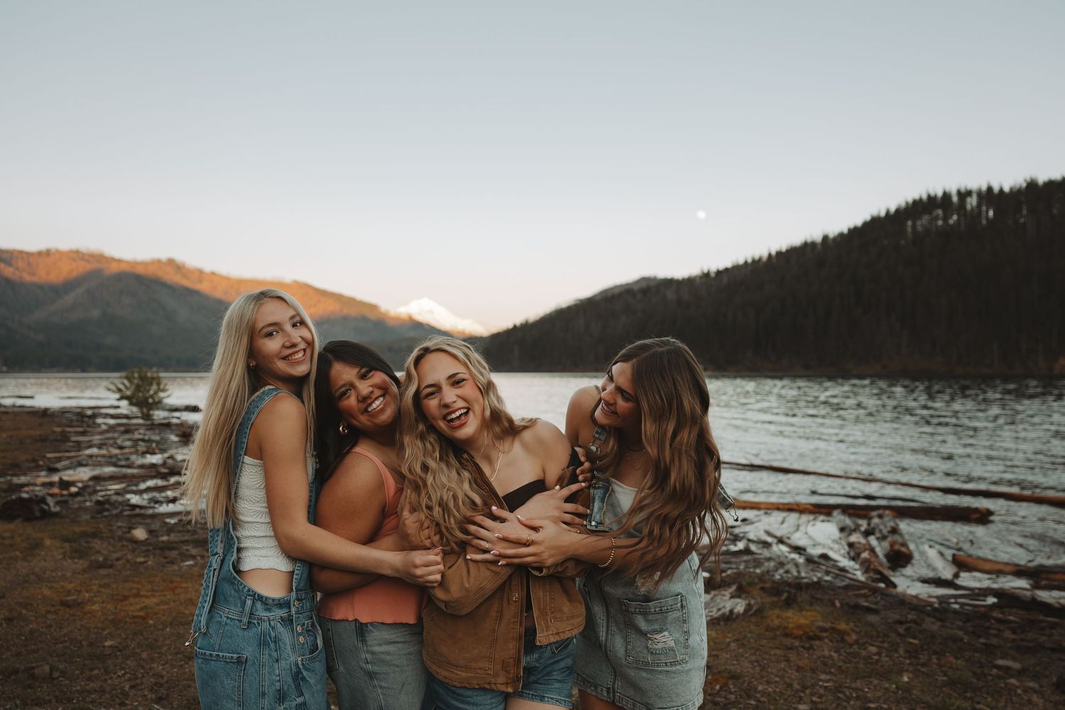Four young women laughing and embracing together on lakeside shore with mountain scenery at sunset.