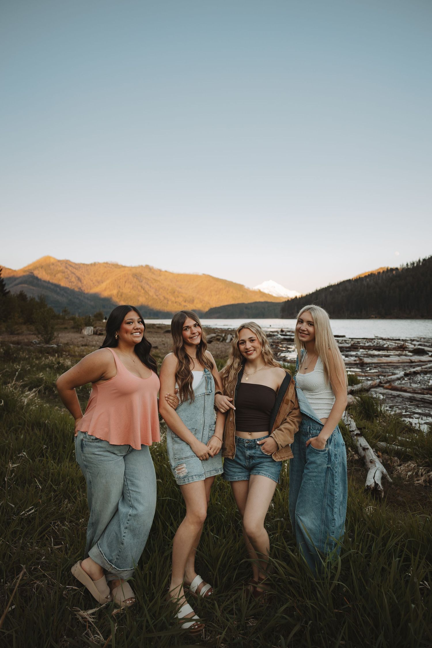 Four women pose together in a grassy field with a mountain lake at golden hour.