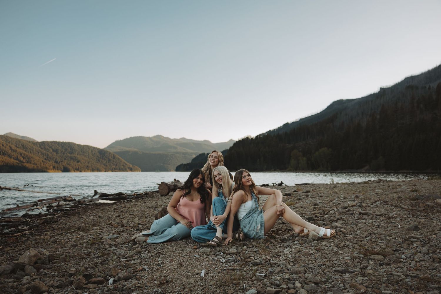 Four young women sitting together on a rocky beach by a mountain lake during golden hour.