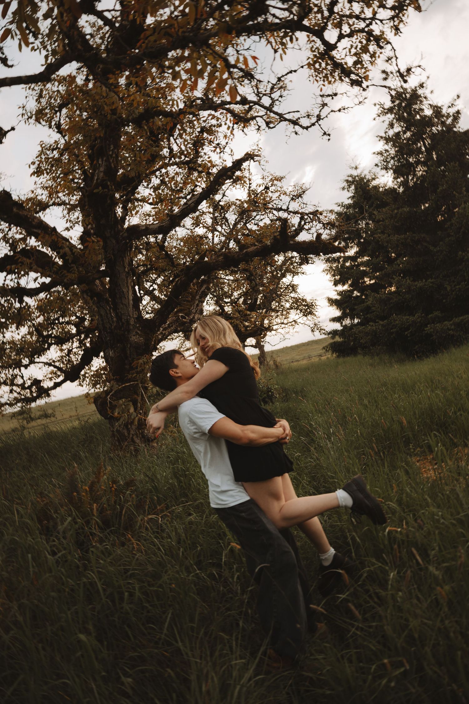 Romantic couple embracing and lifting near a large tree in a lush green meadow at dusk.