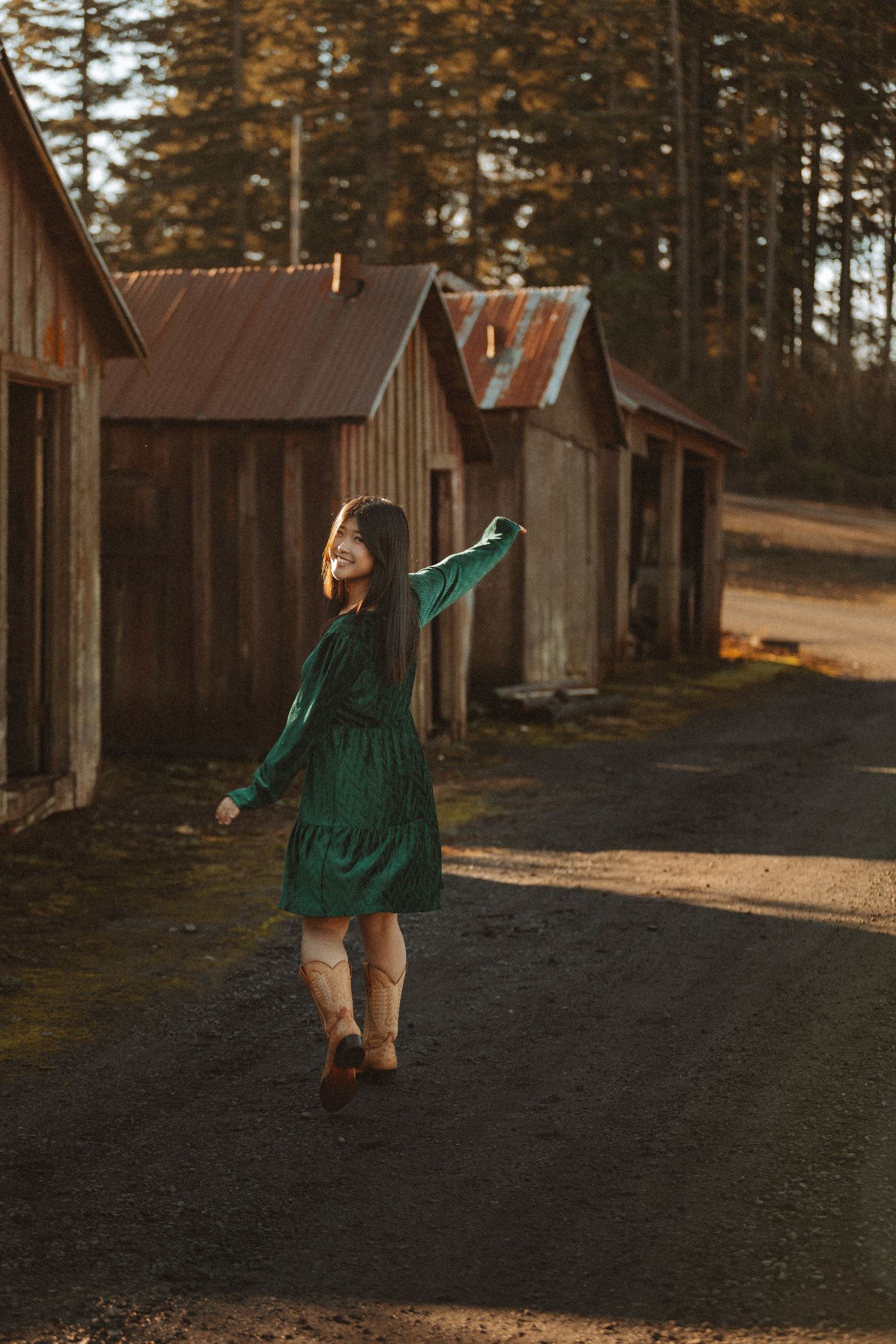 Dancing figure in emerald dress poses joyfully between wooden cabins at sunset.