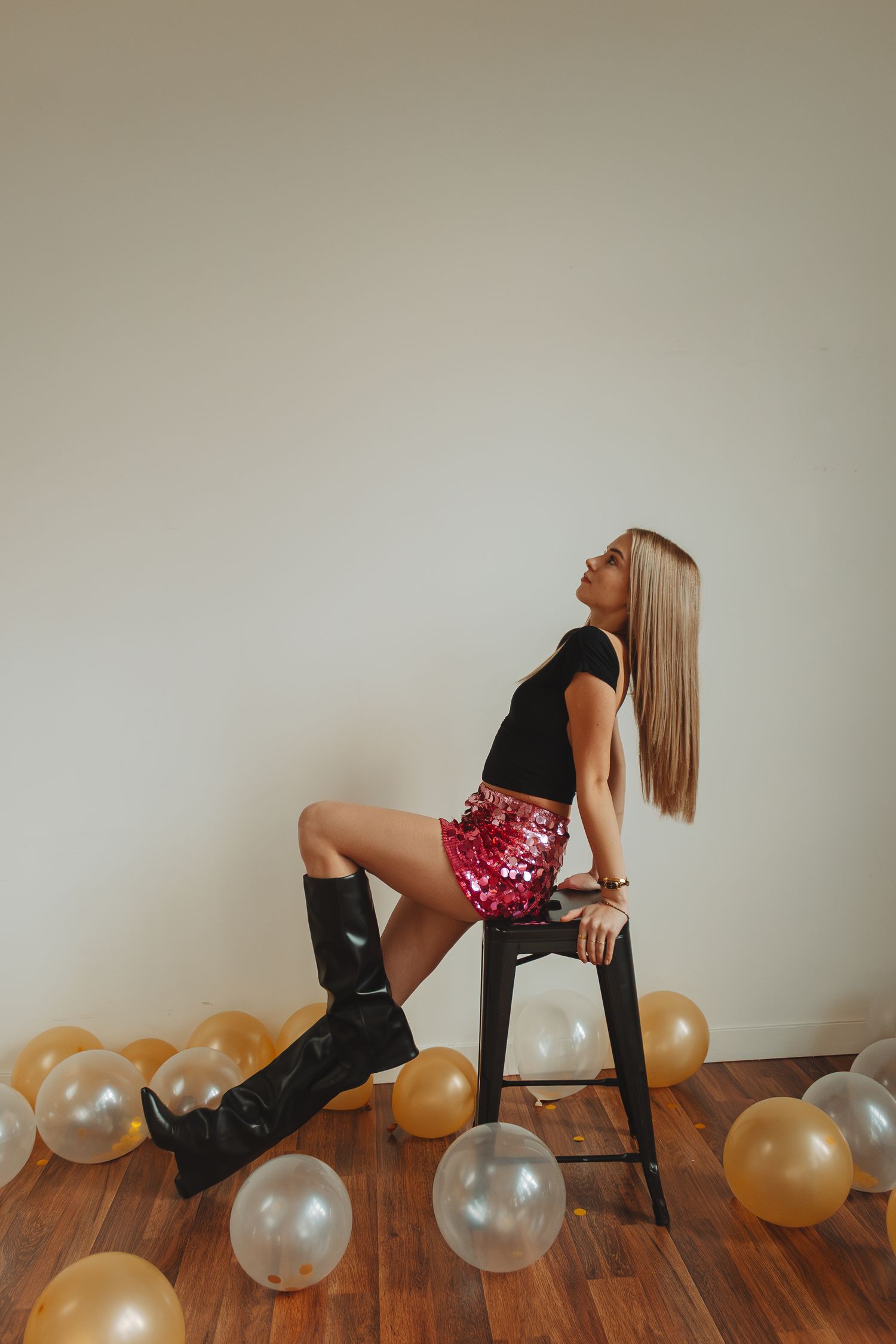 Fashion portrait with gold and white balloons scattered on hardwood floor as someone poses on dark stool.