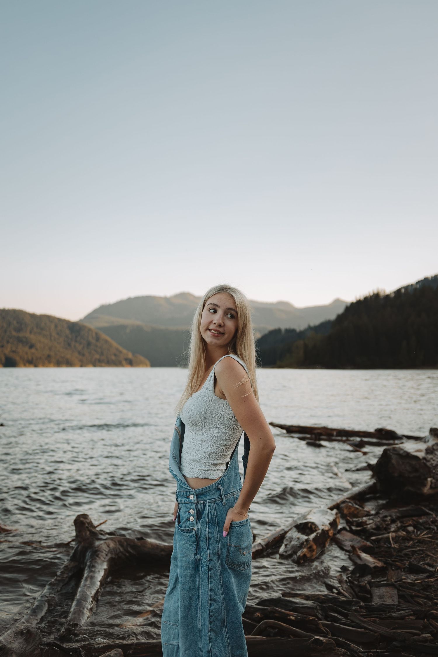 Blonde woman in white tank top and denim skirt standing by a mountain lake with driftwood at dusk.