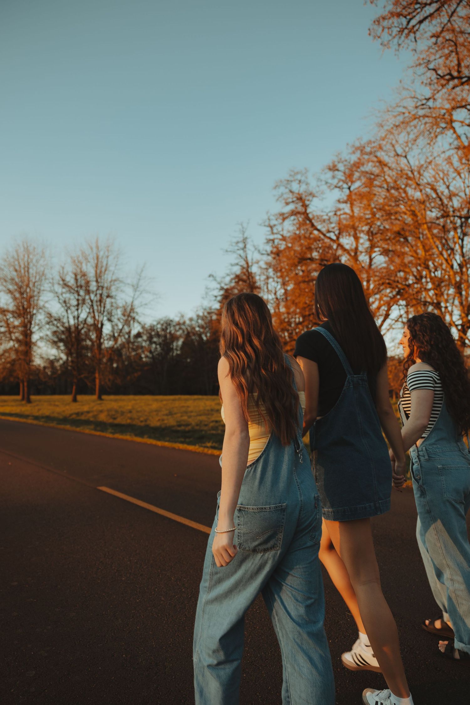 Three girls in denim overalls walk along a tree-lined road at golden hour.