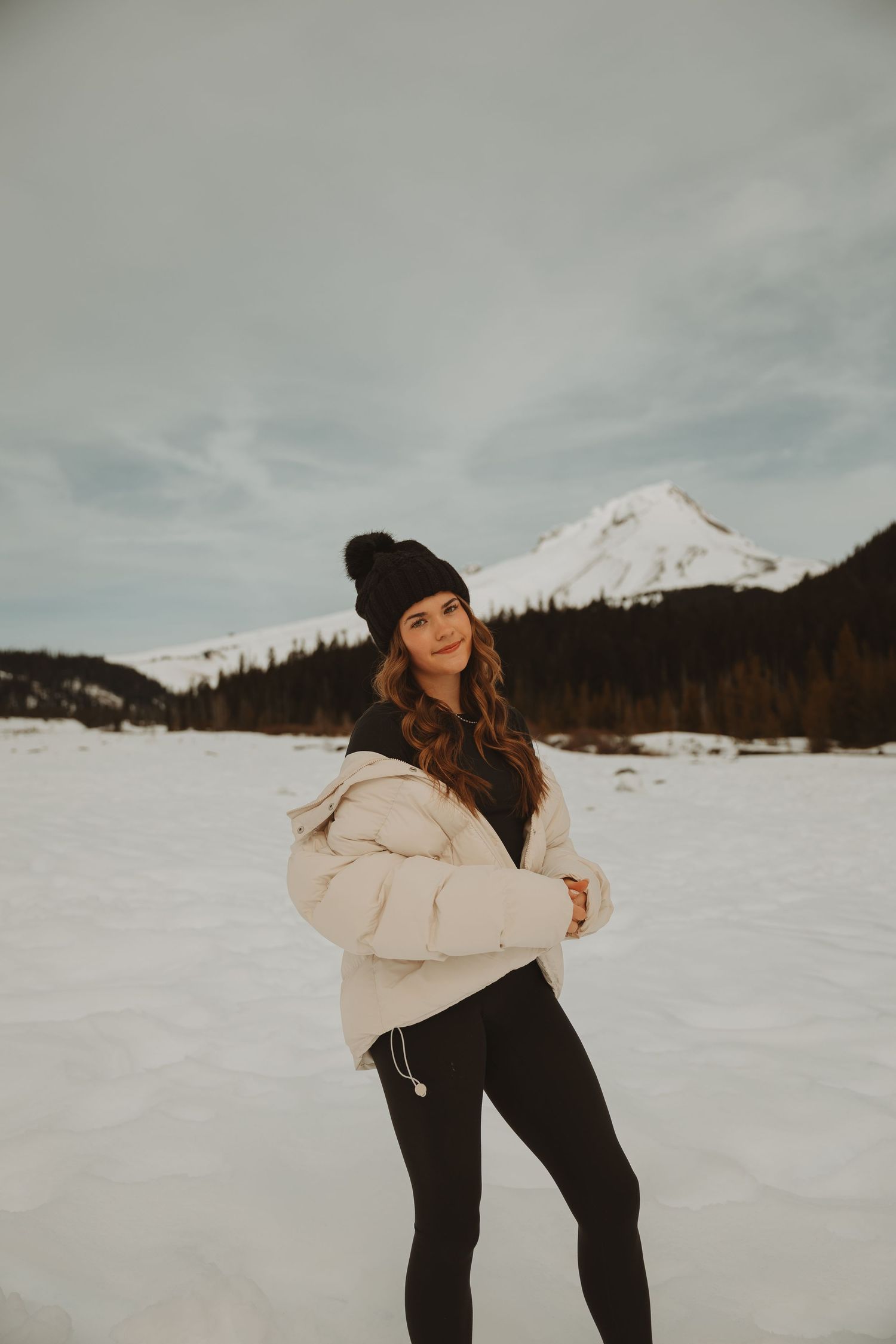 Young woman in white puffer jacket and black beanie poses in snowy mountain landscape with evergreen trees.