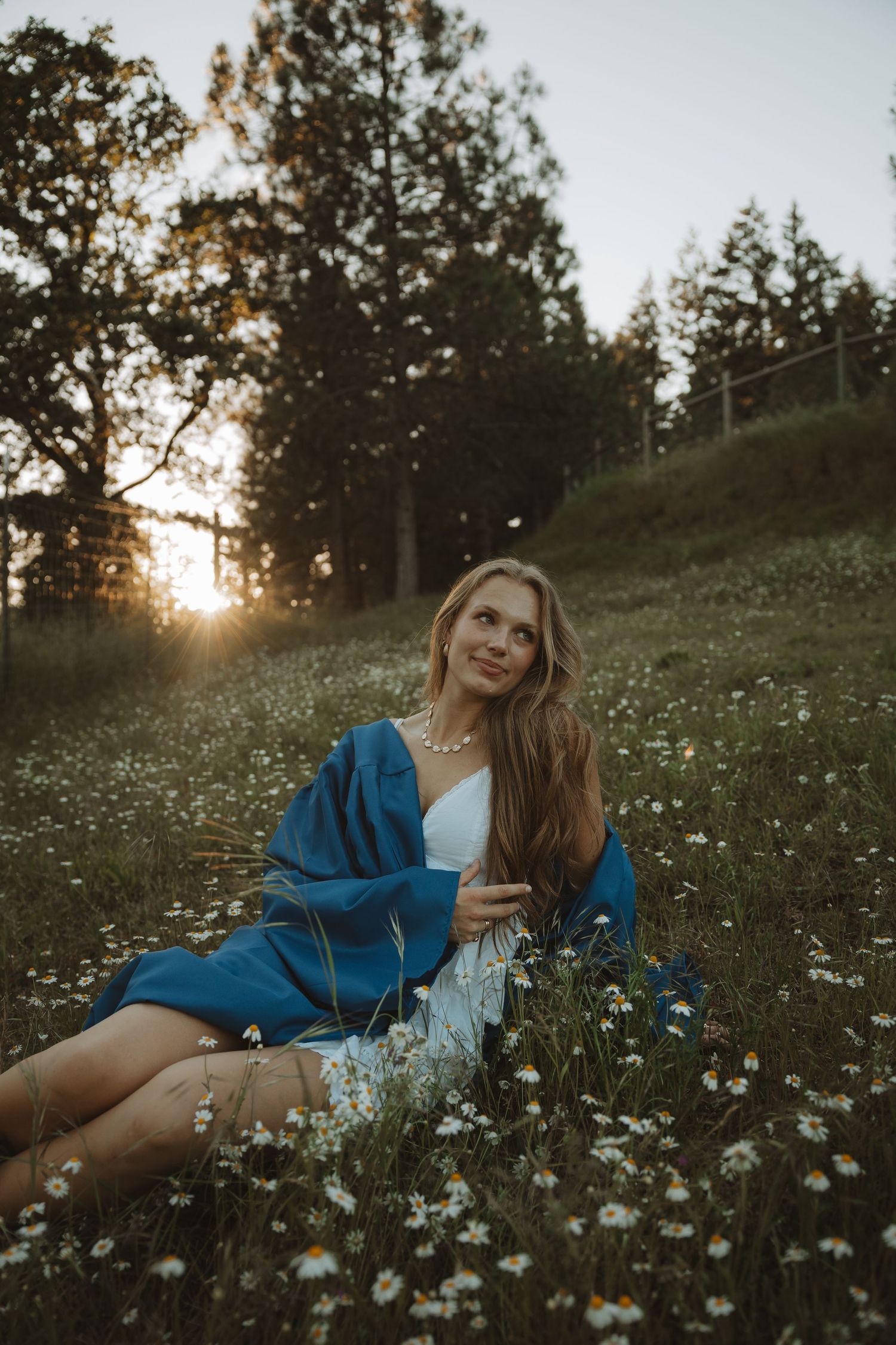 Young woman in blue wrap and white dress sitting in daisy field at golden hour sunset.