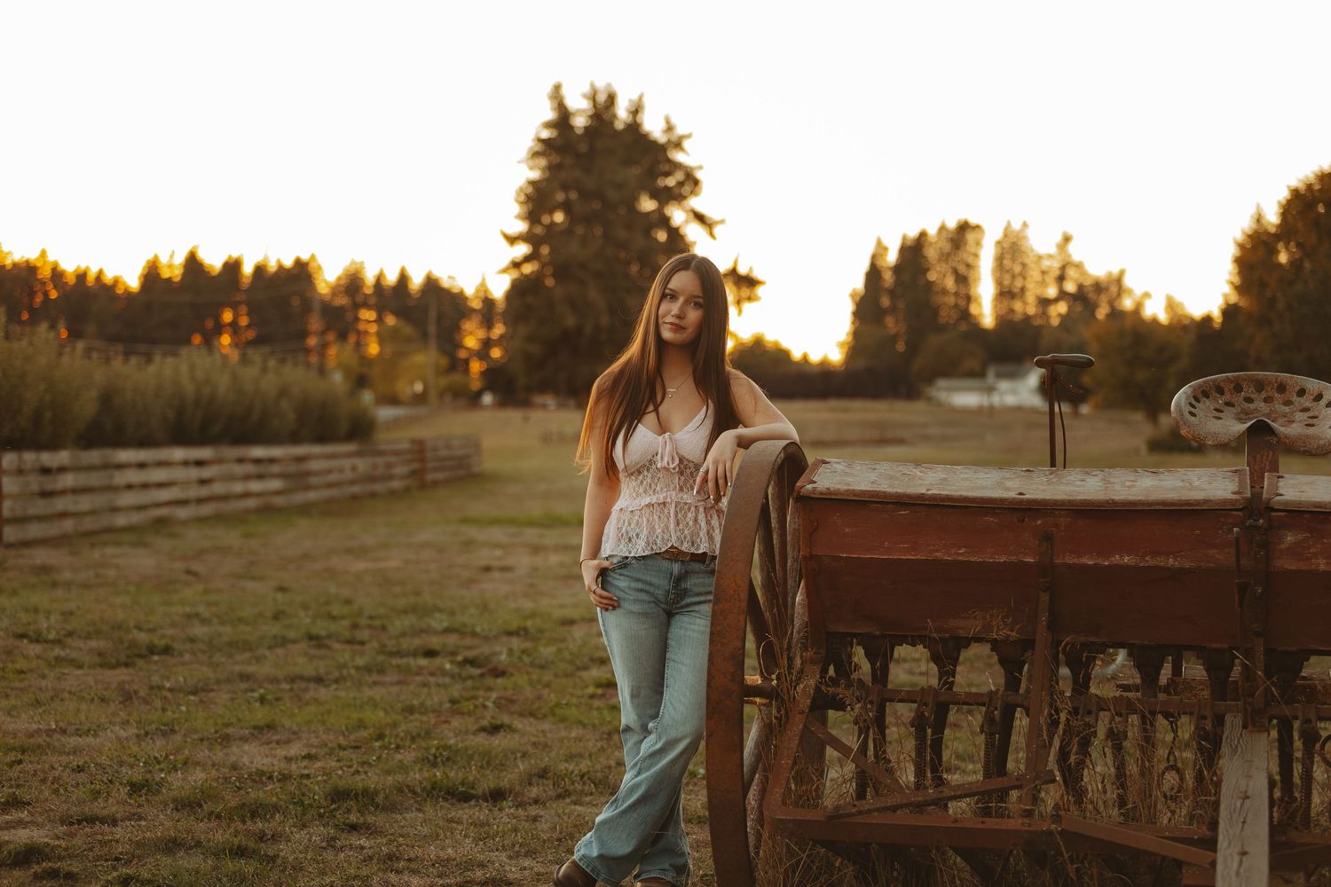 Young woman in jeans and lace top leaning on vintage wooden wagon at golden hour on a rural farm.