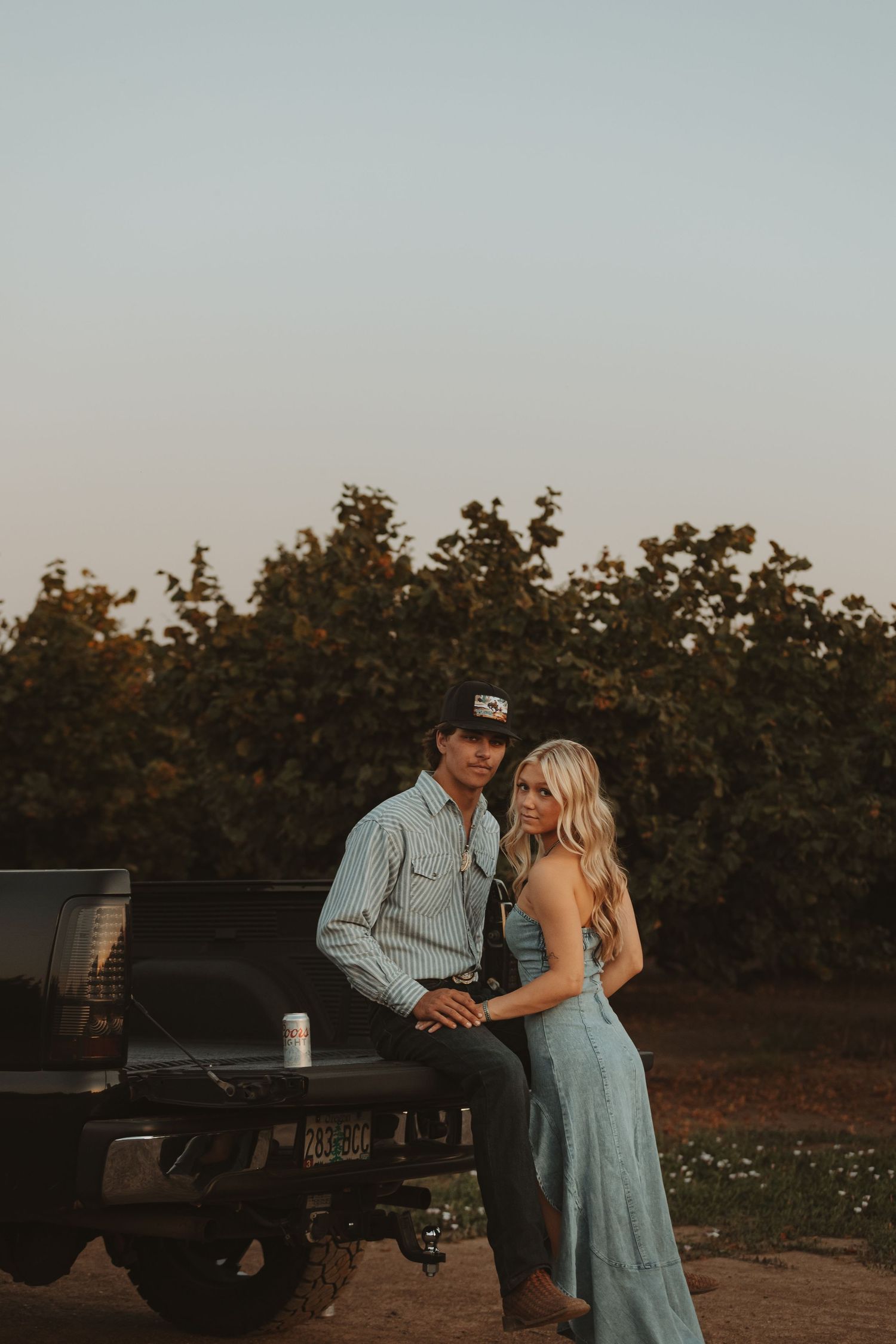 Couple sitting on a truck tailgate at dusk with lush green orchard in the background.
