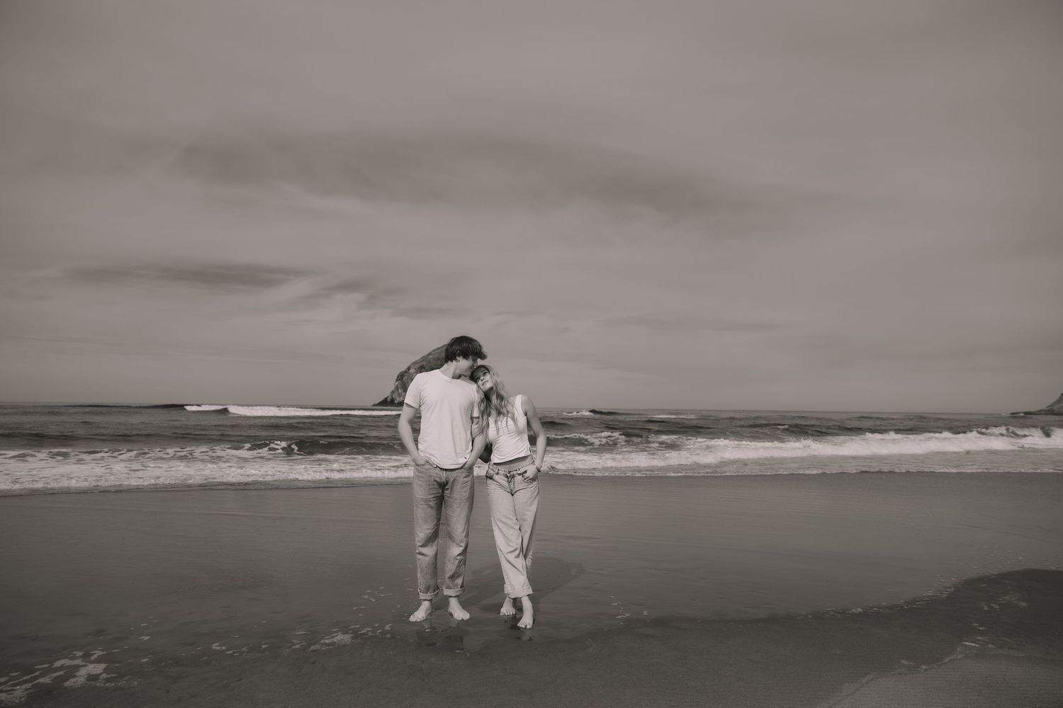 Black and white photo of a couple embracing on a beach with ocean waves and cloudy sky behind them.