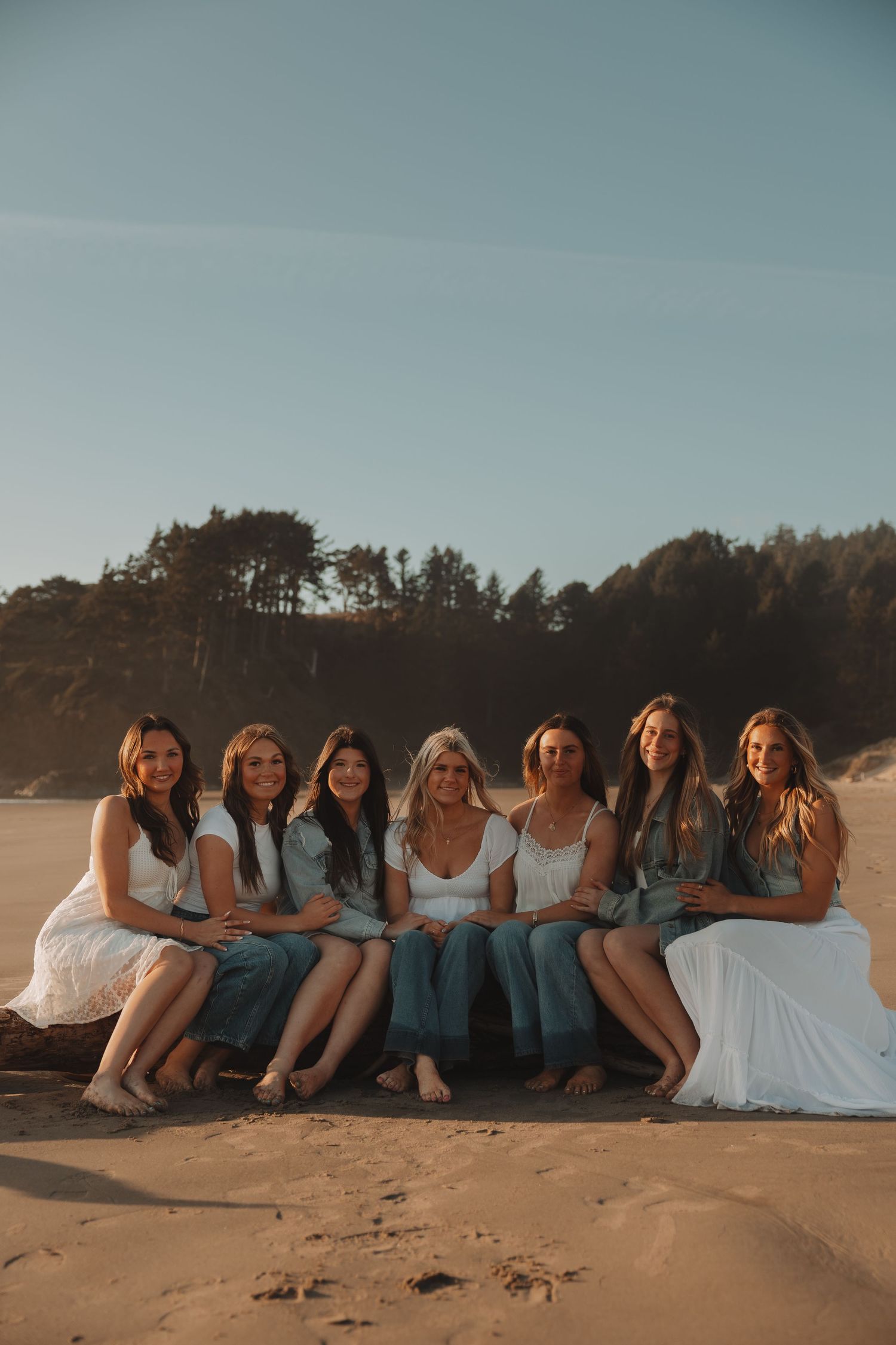 Seven women sit closely together on driftwood on a sandy beach at golden hour.
