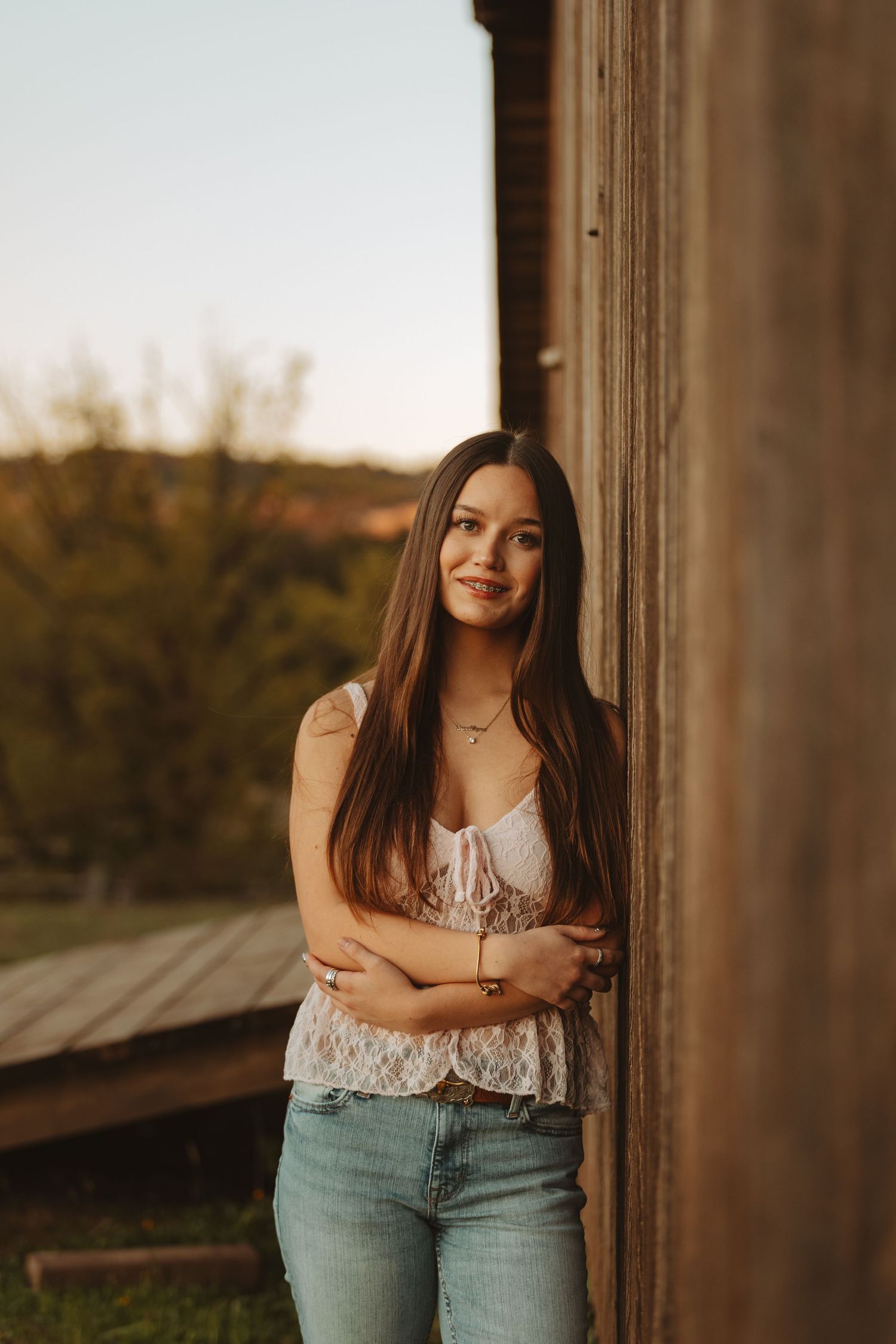 Young woman with long brown hair leans against rustic wooden barn wall in white lace top and jeans at golden hour.