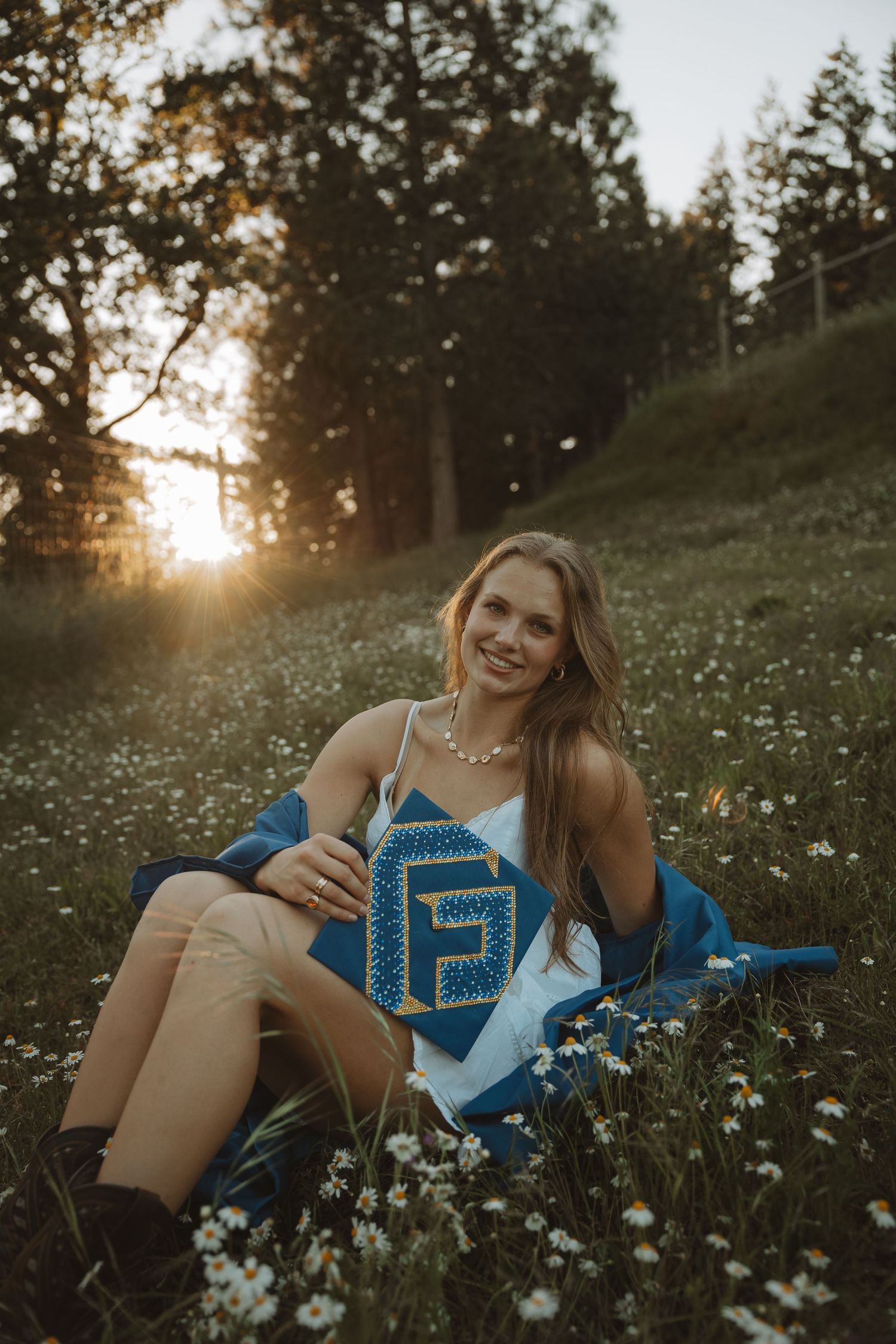 Smiling graduate in blue cap and gown sitting in a wildflower field at golden hour sunset.