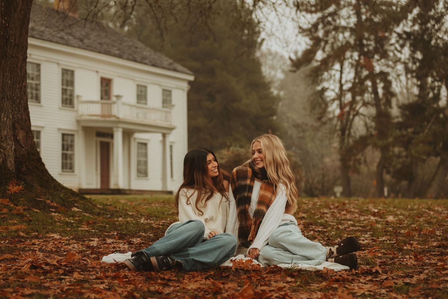 People enjoying a cozy autumn day lounging on blankets among fallen leaves near a white colonial house.