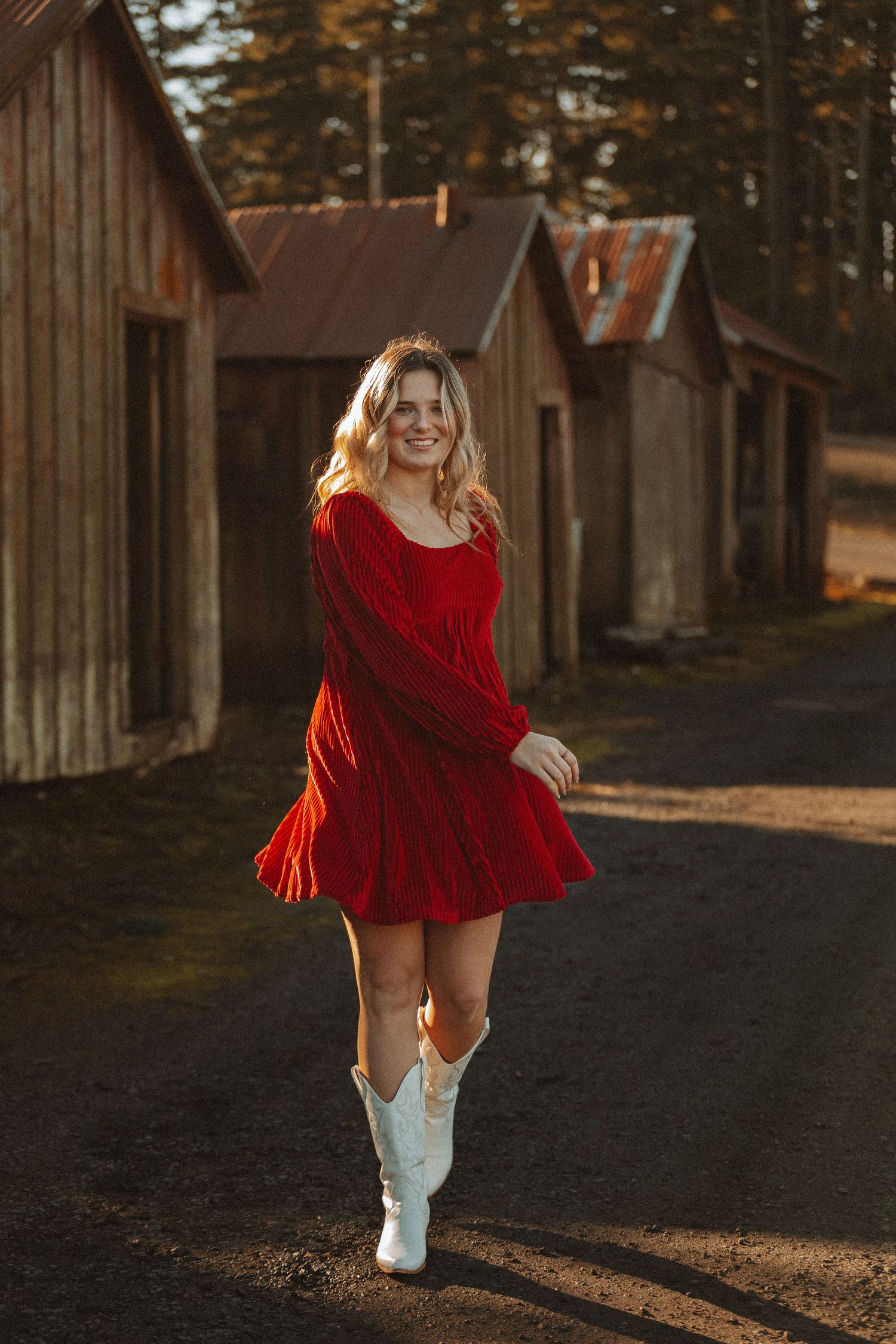A figure in a flowing red dress and white boots dances playfully in front of rustic wooden buildings at sunset.