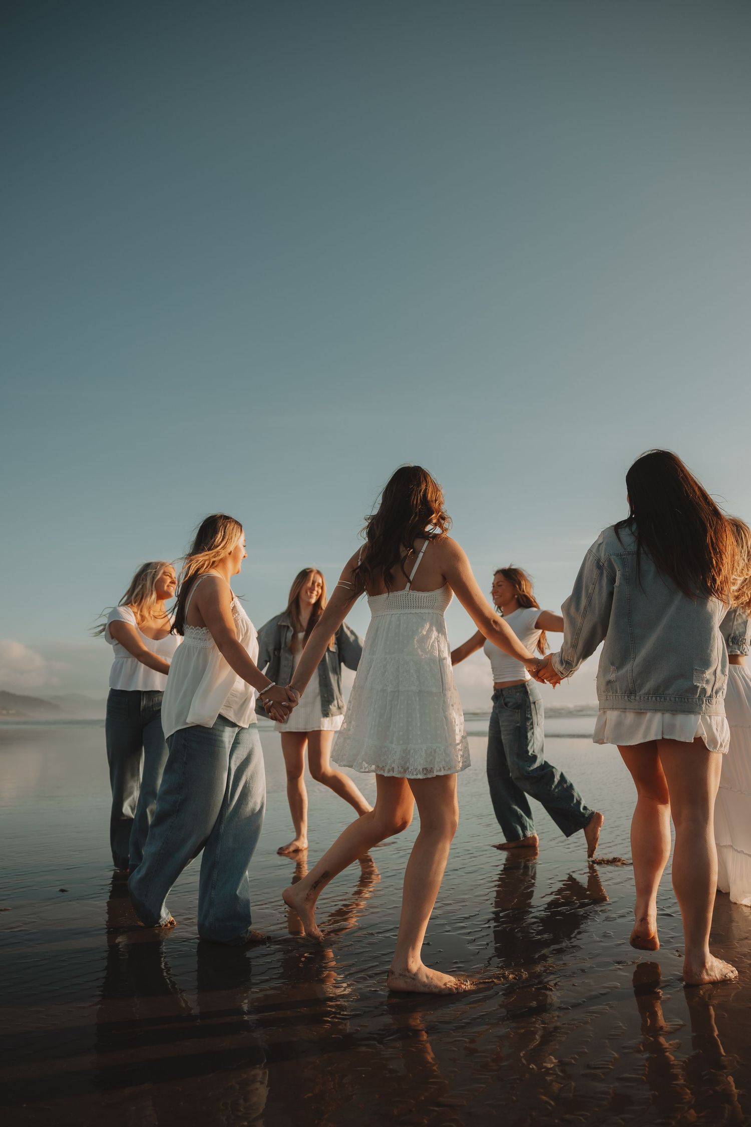 Group of young women dancing in a circle on a reflective wet beach at sunset.
