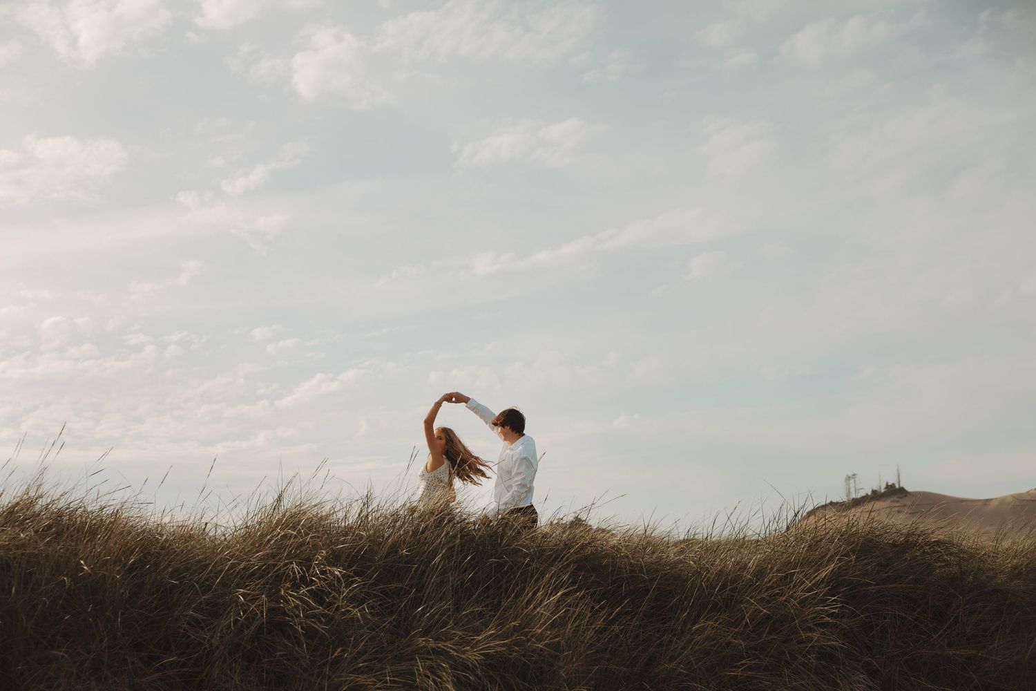 Romantic couple sitting together in tall golden grass field under cloudy sky, holding hands raised above their heads.
