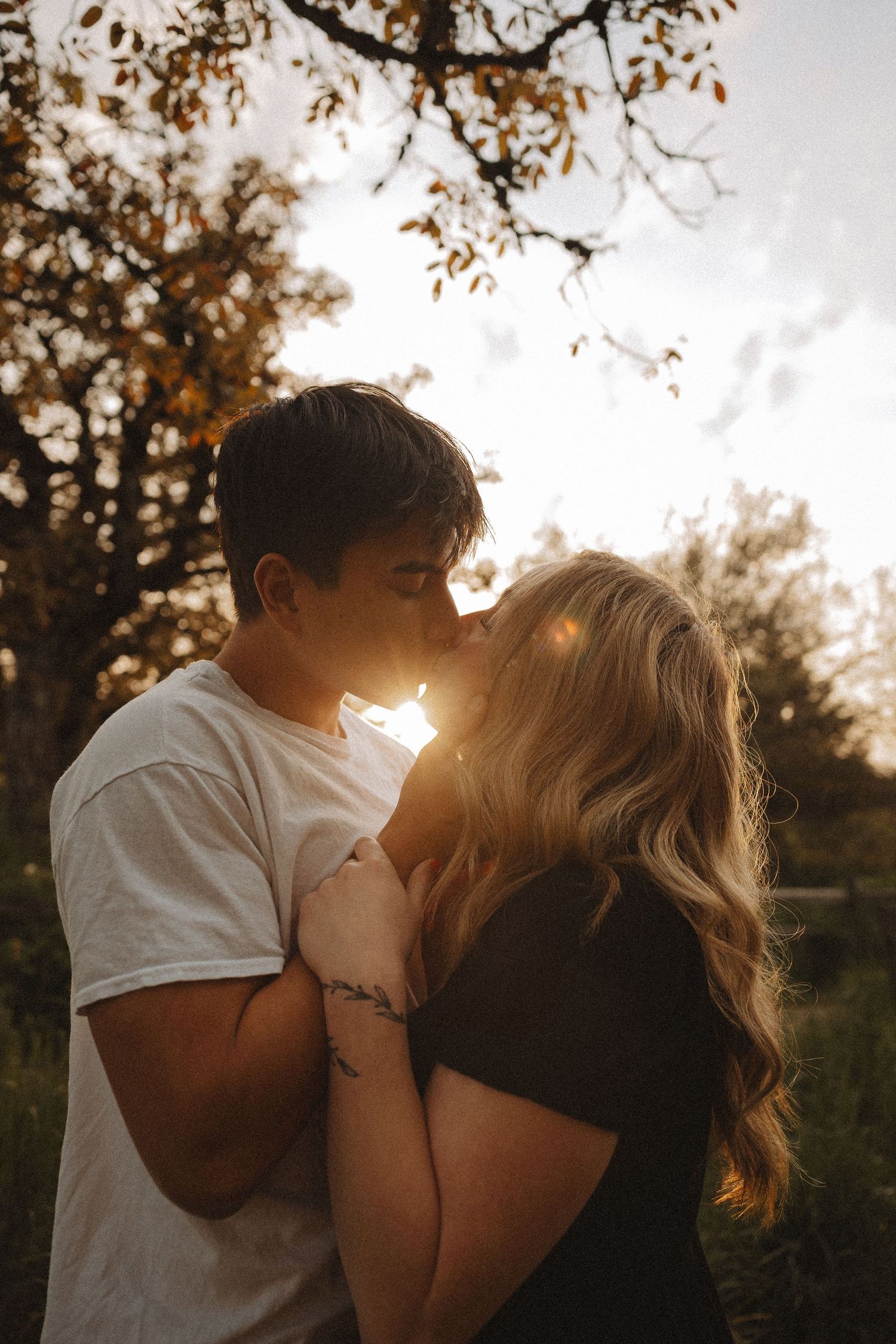 Couple sharing a romantic kiss outdoors at golden sunset, backlit by warm sunlight with autumn trees in background.