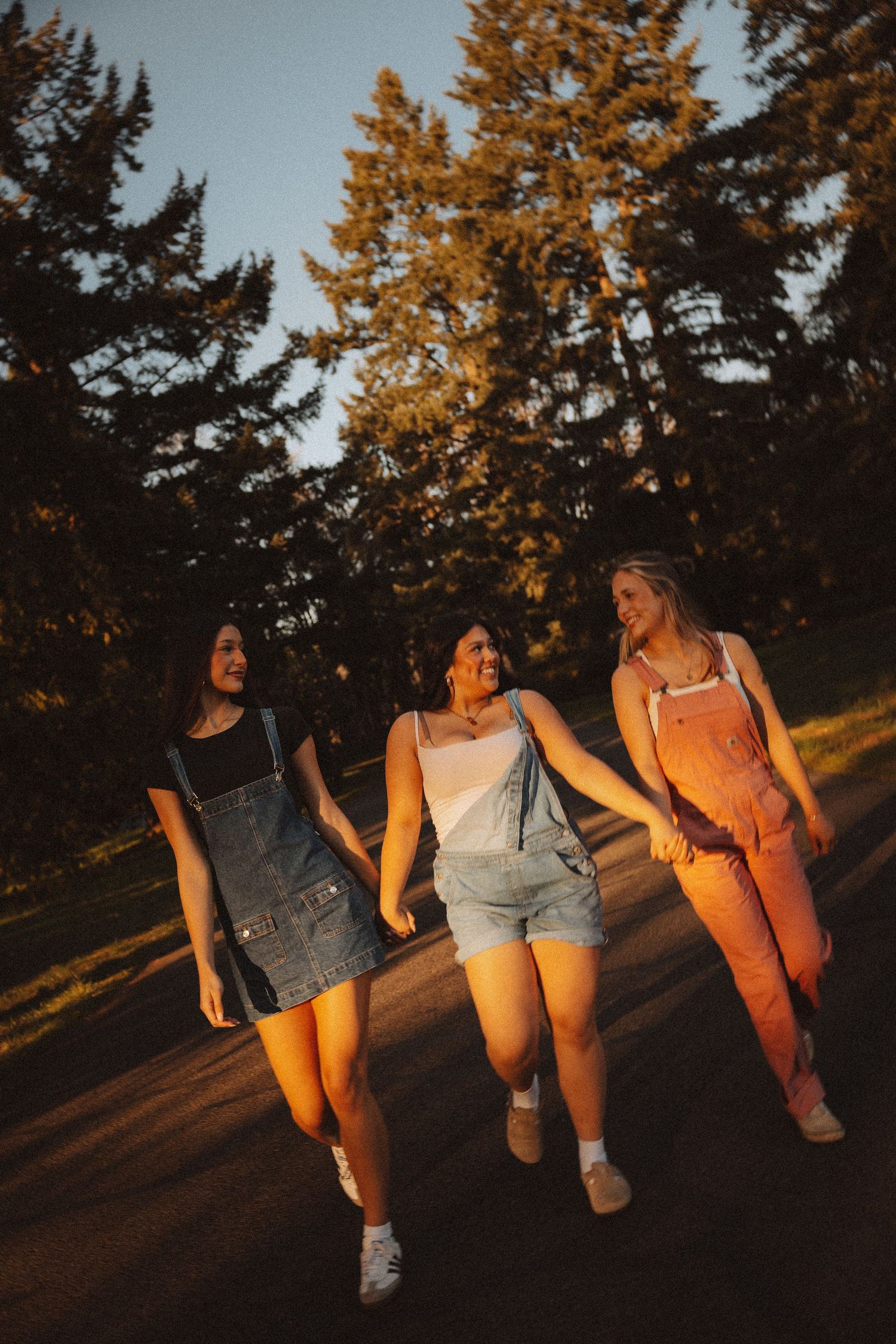 Three young women in overalls walk hand-in-hand down a tree-lined path during golden hour sunset.